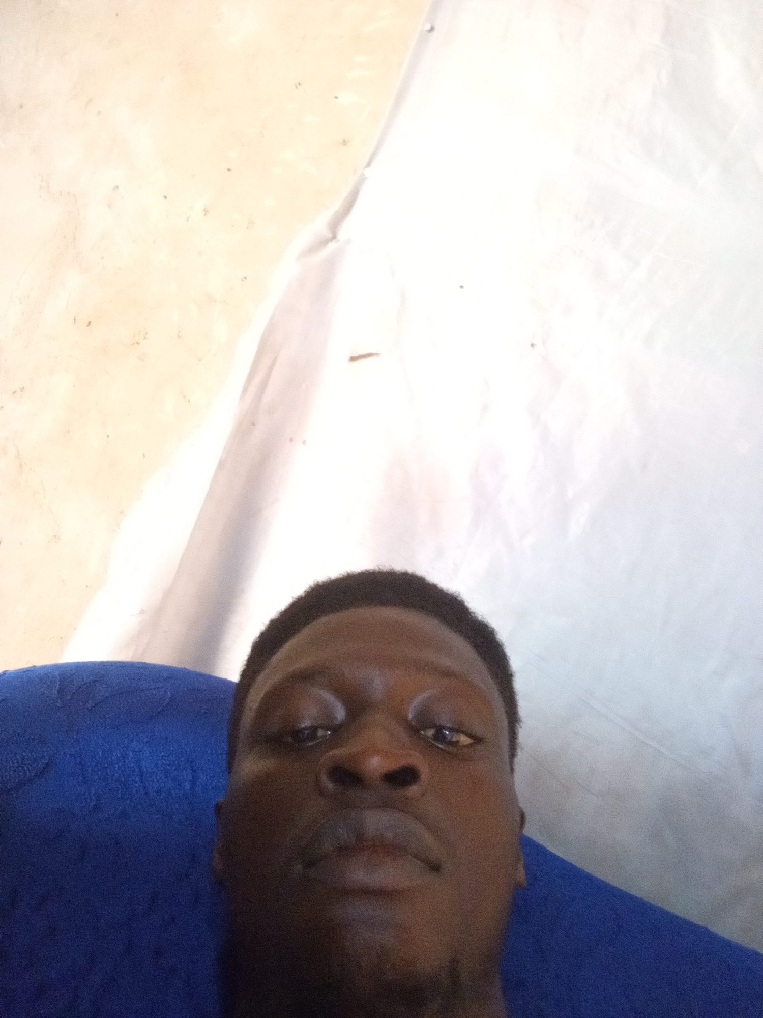 A dark-skinned person is positioned centrally in the frame, looking directly at the camera. Their face occupies the lower portion of the image. A blue textured fabric, possibly upholstery, is visible in the lower left corner. The background consists of a textured, light-colored surface on the left and a large expanse of white, creased material on the right, which could be a sheet or tarp. The environment appears to be indoors, with the lighting suggesting ambient light rather than direct artificial light. There is no specific information in the image to indicate the location within the city of Nyabe, Nigeria.