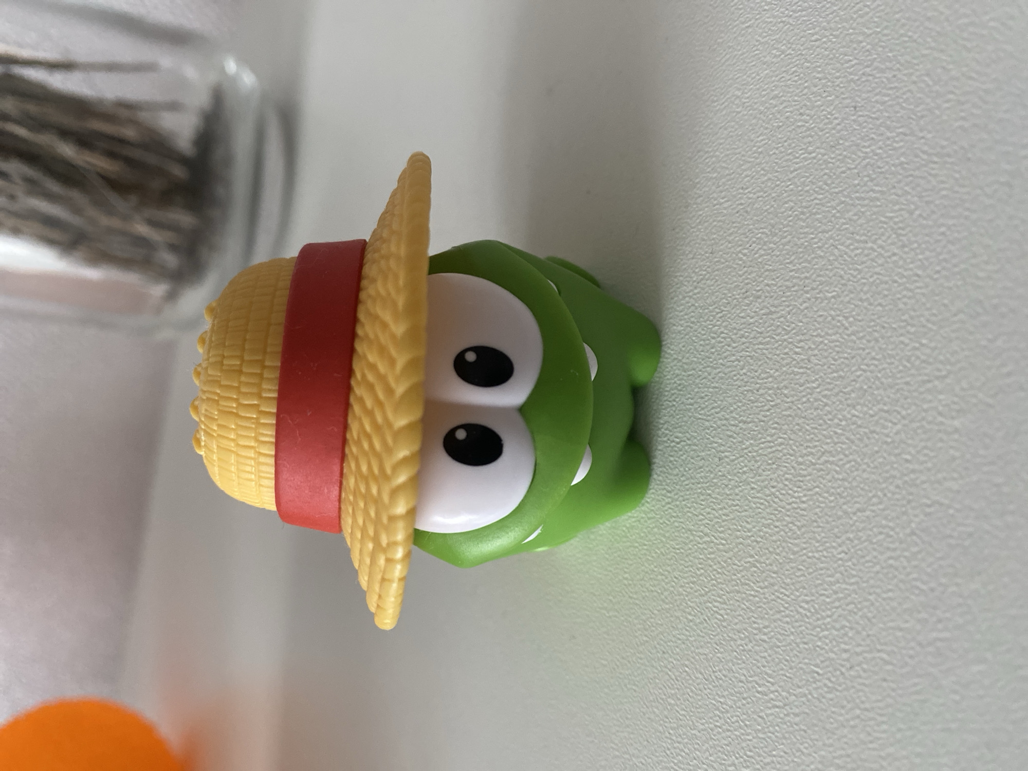 **Location:** Unknown, Unknown.
A small, green Om Nom character toy, wearing a yellow straw hat with a red band, is positioned centrally on a light-colored, textured surface. The toy faces forward, displaying large white eyes with black pupils and two small white fangs protruding from its lower face. In the upper left background, a blurred orange spherical object is partially visible. To the upper right background, a blurred clear glass container filled with dark, dried plant material is present. The scene is evenly lit.