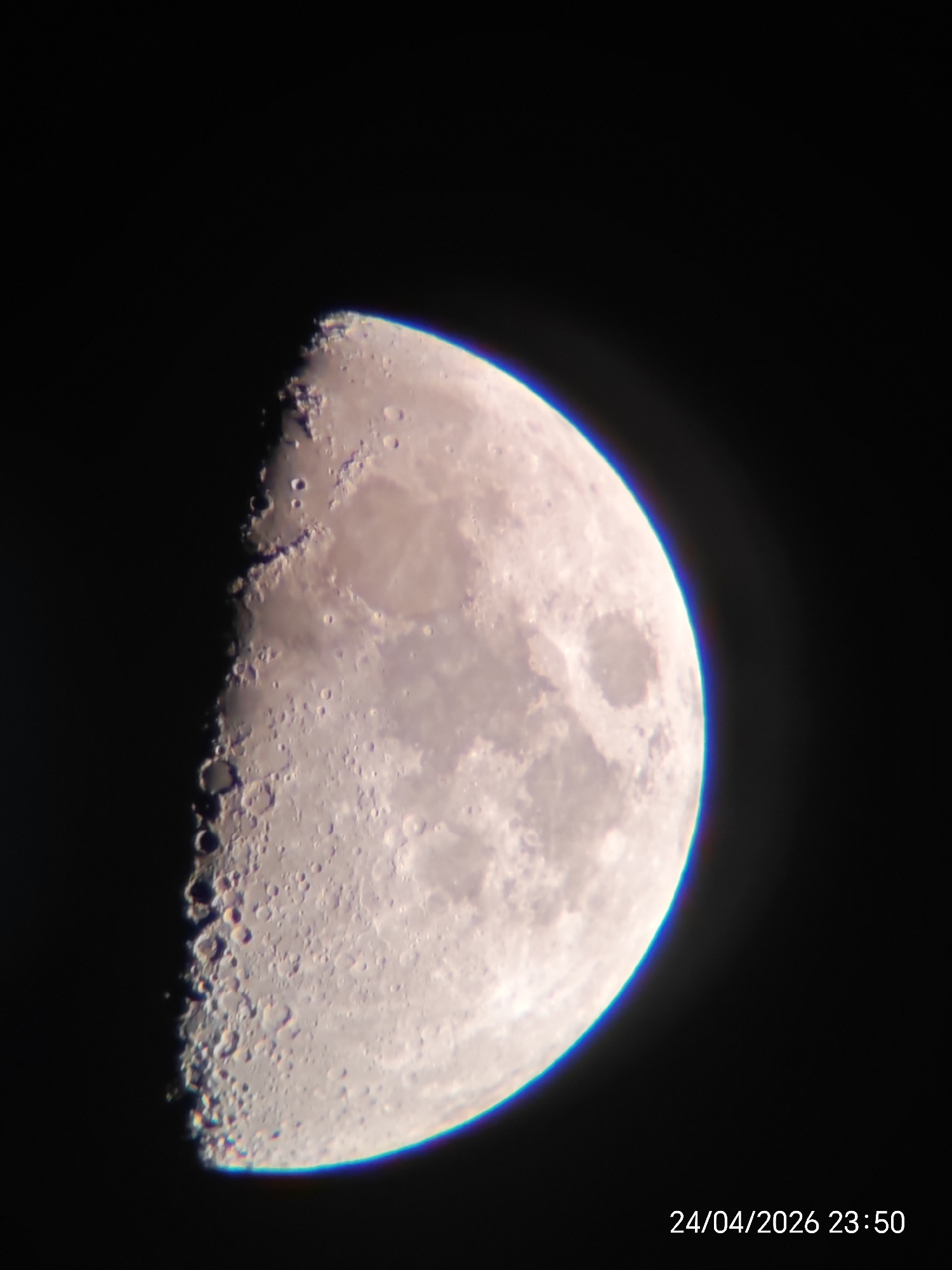 This media file displays an astronomical photograph of the Moon, prominently featured against a dark, black background representing outer space. The Moon is in a gibbous phase, with more than half of its surface illuminated, showing distinct surface features.
The illuminated portion of the Moon reveals numerous impact craters of varying sizes, particularly concentrated along the terminator – the jagged line separating light and shadow. Mountains and rough terrain are also visible along this line, casting long shadows that accentuate their relief. Smoother, darker regions, likely lunar maria, contrast with the brighter, heavily cratered highlands.
A noticeable chromatic aberration, appearing as a subtle blue or purple fringe, is visible along the illuminated limb of the Moon, especially on the right side. The Moon's overall color palette consists of light grey-white tones with faint hints of pale yellow or brown in certain areas.
In the bottom right corner of the main image, white text is visible, stating "24/04/2026 23:50," indicating the date and time the photograph was captured. The image captures a detailed view of the Moon's topography through astrophotography. No people or specific terrestrial location cues are present.