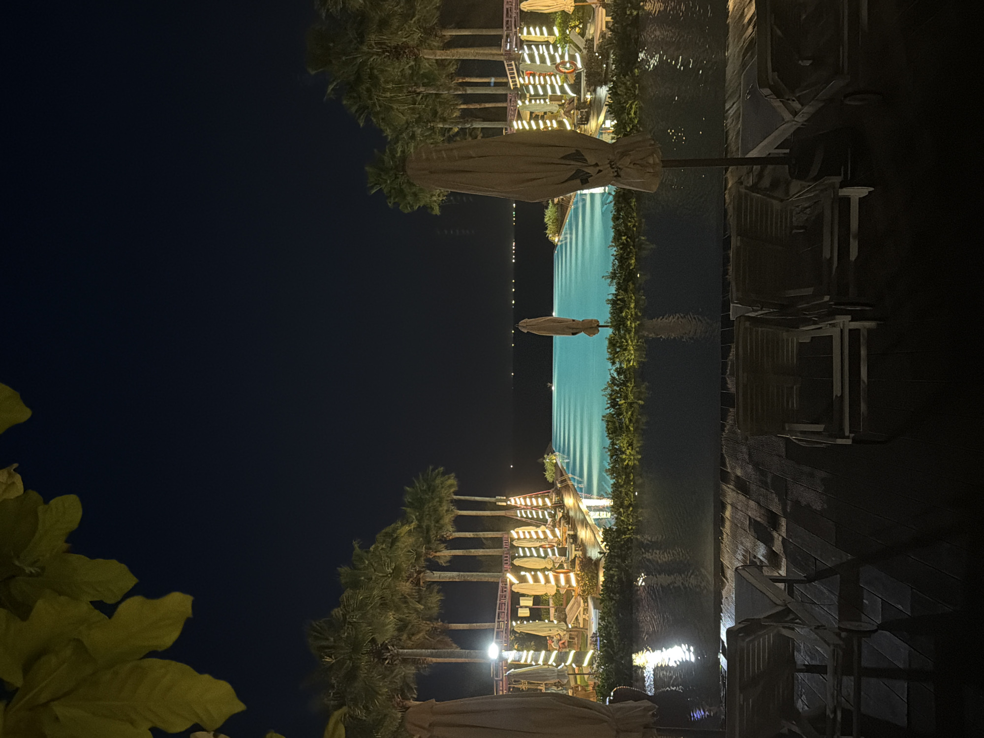 This image depicts a resort at night in Dong Ninh Hoa Ward, Vietnam. The scene is dominated by a large, illuminated turquoise infinity pool extending towards a dark body of water on the horizon, where distant lights are visible. On either side of the pool, multiple tall palm trees stand, interspersed with structures and closed beige umbrellas wrapped with string lights. In the immediate foreground, a dark wooden deck holds three empty wooden lounge chairs. A shallow, reflective water feature separates the deck from the main infinity pool. Green foliage frames the lower pool edge and the upper left corner of the image. No individuals are present.