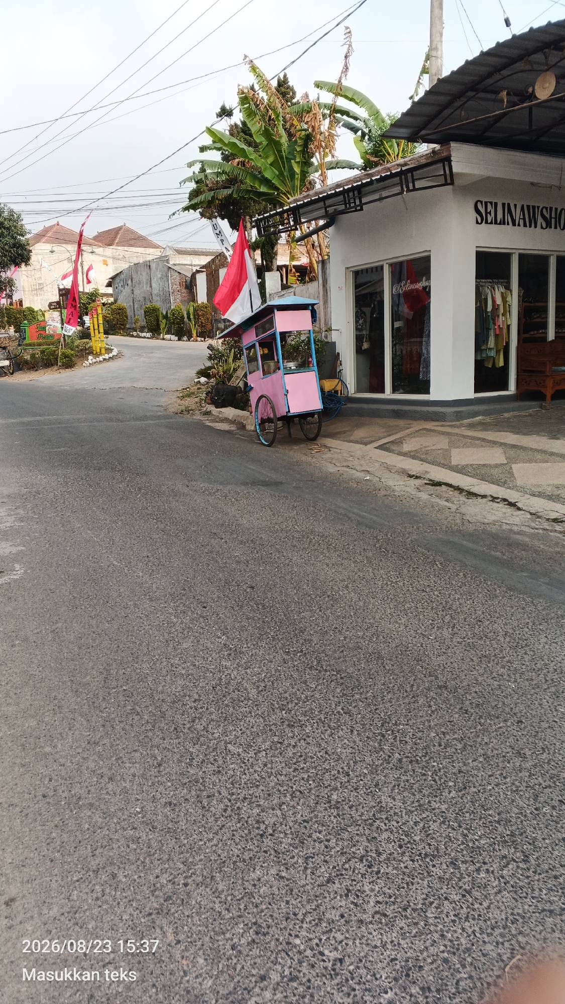 In the city of Jeru, Indonesia, a pink and blue food cart is parked on the side of a paved road. The cart is adorned with two Indonesian flags. A white building with the text "SELINAWSHOP" is visible behind the cart. The environment appears to be a street scene with buildings and trees in the background. The sky is overcast. A date and time stamp "2026/08/23 15:37" and text "Masukkan teks" are present in the bottom left corner of the image.