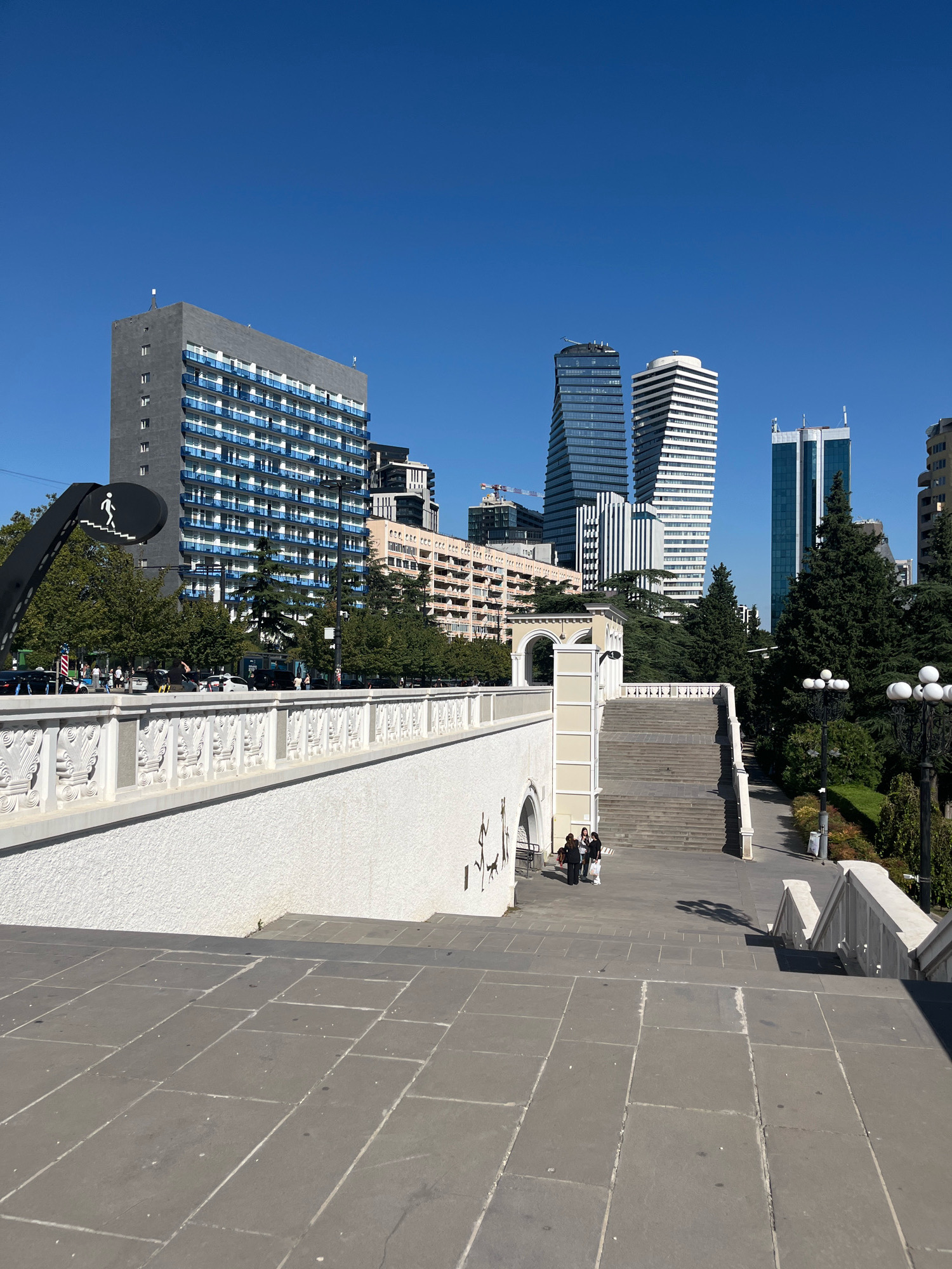 The image depicts a bright, sunny day in Tbilisi, Georgia, with a clear blue sky. In the foreground, a wide, grey stone staircase descends, bordered by white balustrades adorned with decorative carvings. The balustrades continue along a white, textured wall to the left. Further up the stairs, a small group of three people, two women and a man, are walking. To the left of the staircase, a large, black abstract sculpture of a figure walking down stairs is visible. Beyond the staircase and decorative wall, the cityscape of Tbilisi is laid out. Several modern skyscrapers, characterized by glass facades and geometric designs, dominate the background. Among them are a dark blue glass tower, two white and black striped towers, and a lighter blue glass tower. In the mid-ground, there are trees and a multi-story building with a facade of balconies. The overall scene is urban and modern, with a blend of architectural styles.