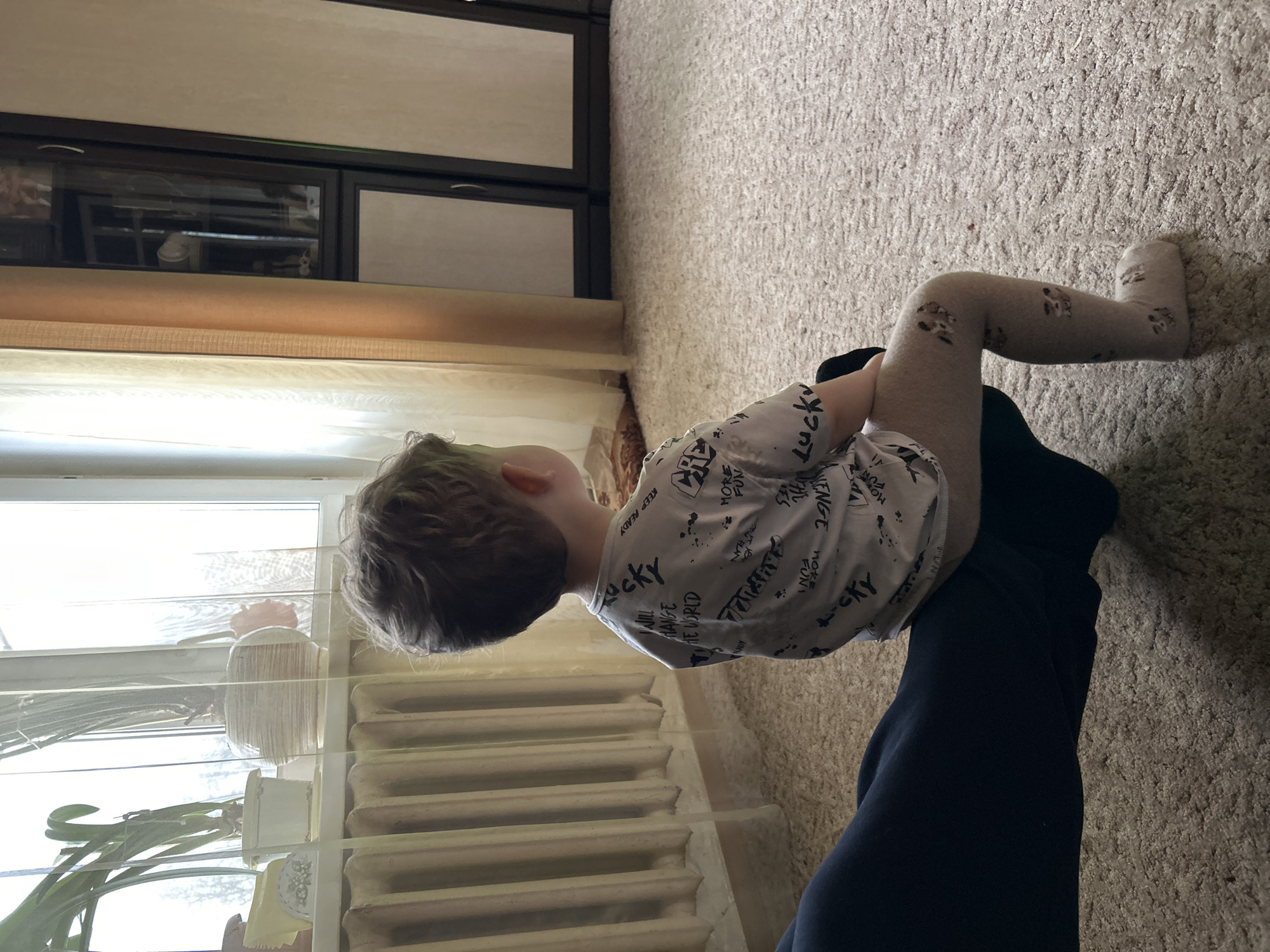 A child in patterned tights is being held by an adult in a residential room. The child is wearing a t-shirt with printed words, including "lucky". The child's legs extend onto a carpeted floor. A radiator sits near the child. There is a window with blinds and plants to the left. A dark wooden cabinet is in the background. Given the provided city and country, the scene is located in a home in Klin, Russia.