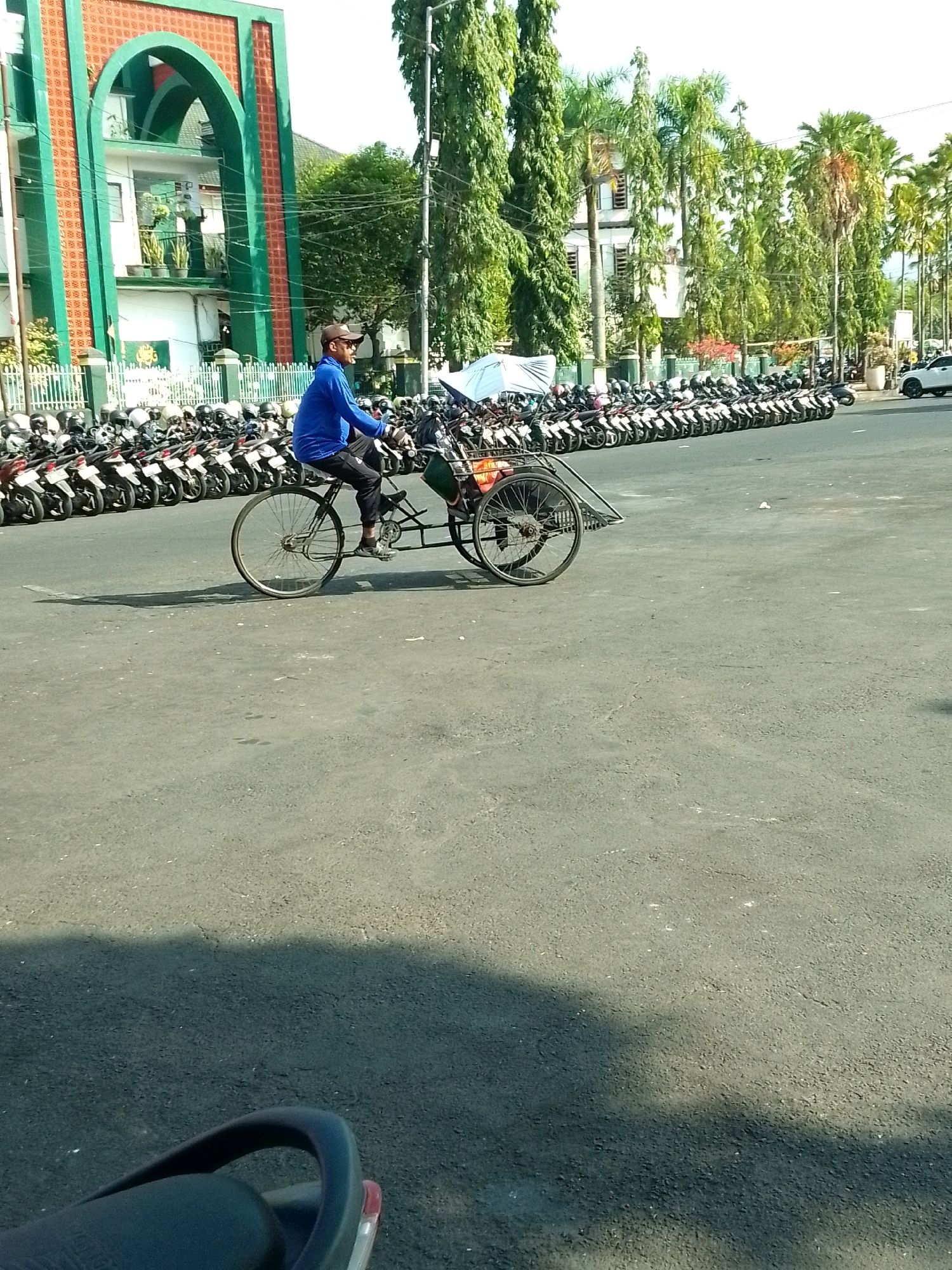 The image depicts a street scene in Jember, Indonesia, near a building with a distinctive green archway and brick accents, likely a mosque or similar institution. In the foreground, a man wearing a blue long-sleeved shirt, dark pants, and a baseball cap is riding a three-wheeled bicycle-powered rickshaw. He is looking forward and appears to be in motion. The rickshaw has a large spoked wheel at the back and two smaller wheels in the front, with a basket-like cargo area behind the rider.
Behind the rickshaw, a vast number of motorcycles are parked in neat rows, suggesting a public area or event. The background features tall, slender trees, characteristic of tropical climates, and a white building. The ground is paved asphalt, and shadows are cast by the sun, indicating it is daytime. The overall atmosphere is that of a busy, sunny day in an urban Indonesian setting.