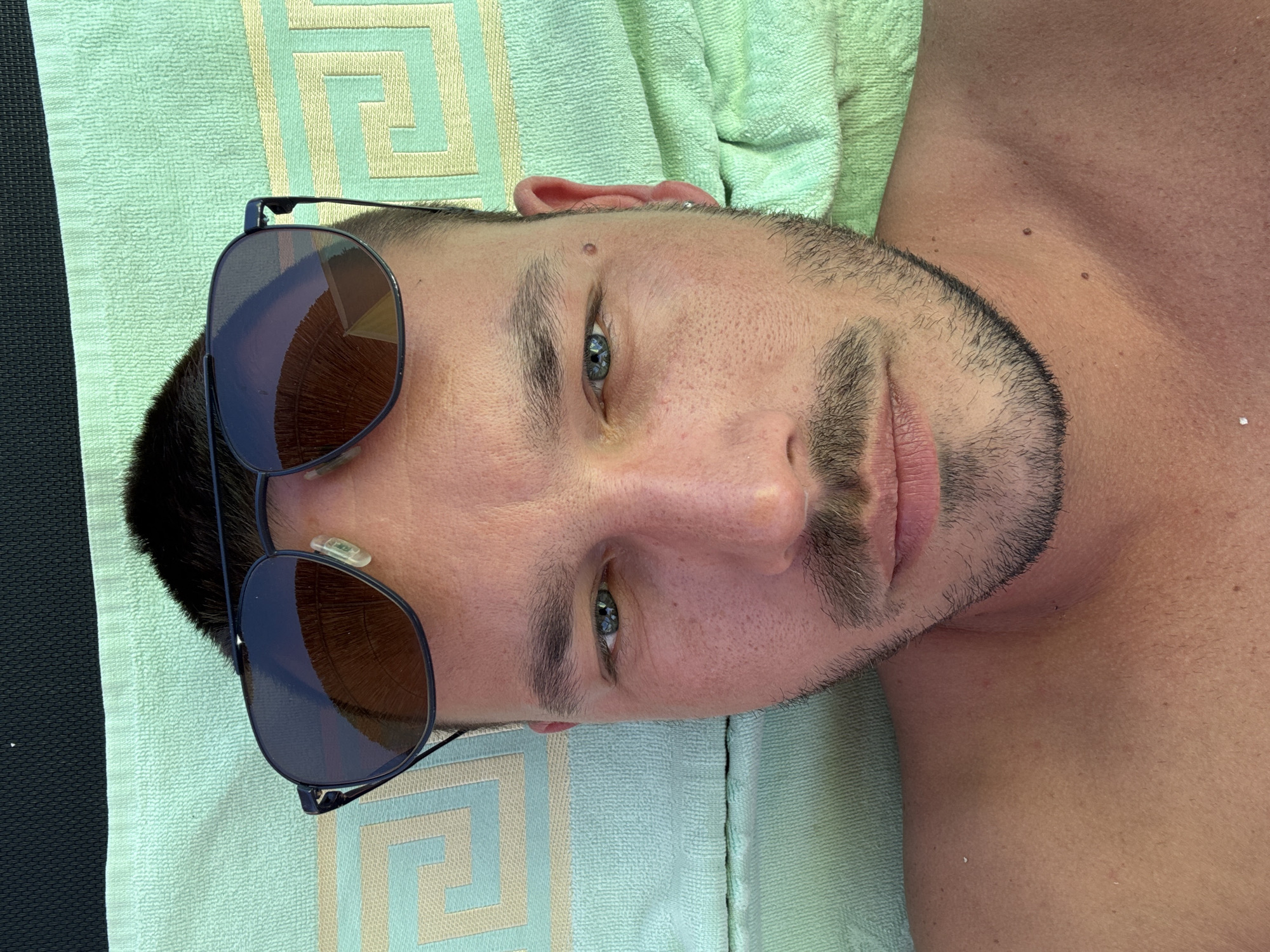 A man, wearing sunglasses, is lying down on a light green towel with a patterned border. He has short dark hair and a mustache and beard. The towel is on a surface that appears to be a beach chair or lounger, indicated by a dark fabric at the top of the image. The man is looking directly at the camera. The setting is likely outdoors, possibly a beach or poolside area, in Toroni Municipal Unit, Greece, given the presence of sun and beach attire.