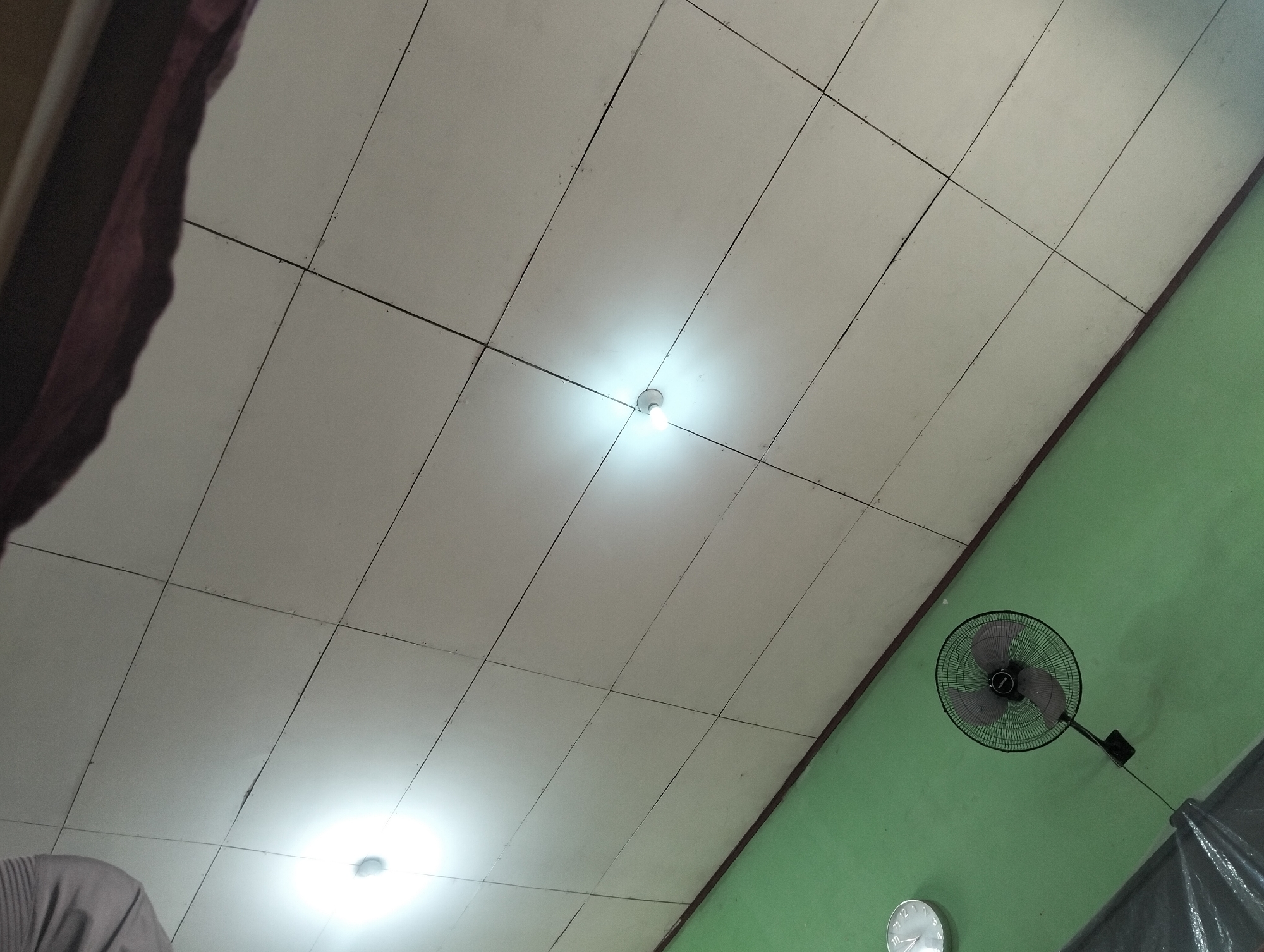 The image shows a room with a tiled ceiling and a green wall. A ceiling fan is mounted on the green wall, and a clock is visible below it. There are two light bulbs on the ceiling, one of which is illuminated. A portion of a person's shoulder and arm wearing a grey garment is visible in the lower left corner of the image. The scene appears to be indoors in a residential or office setting. The specific location in Banjar Agung, Indonesia, is unknown.