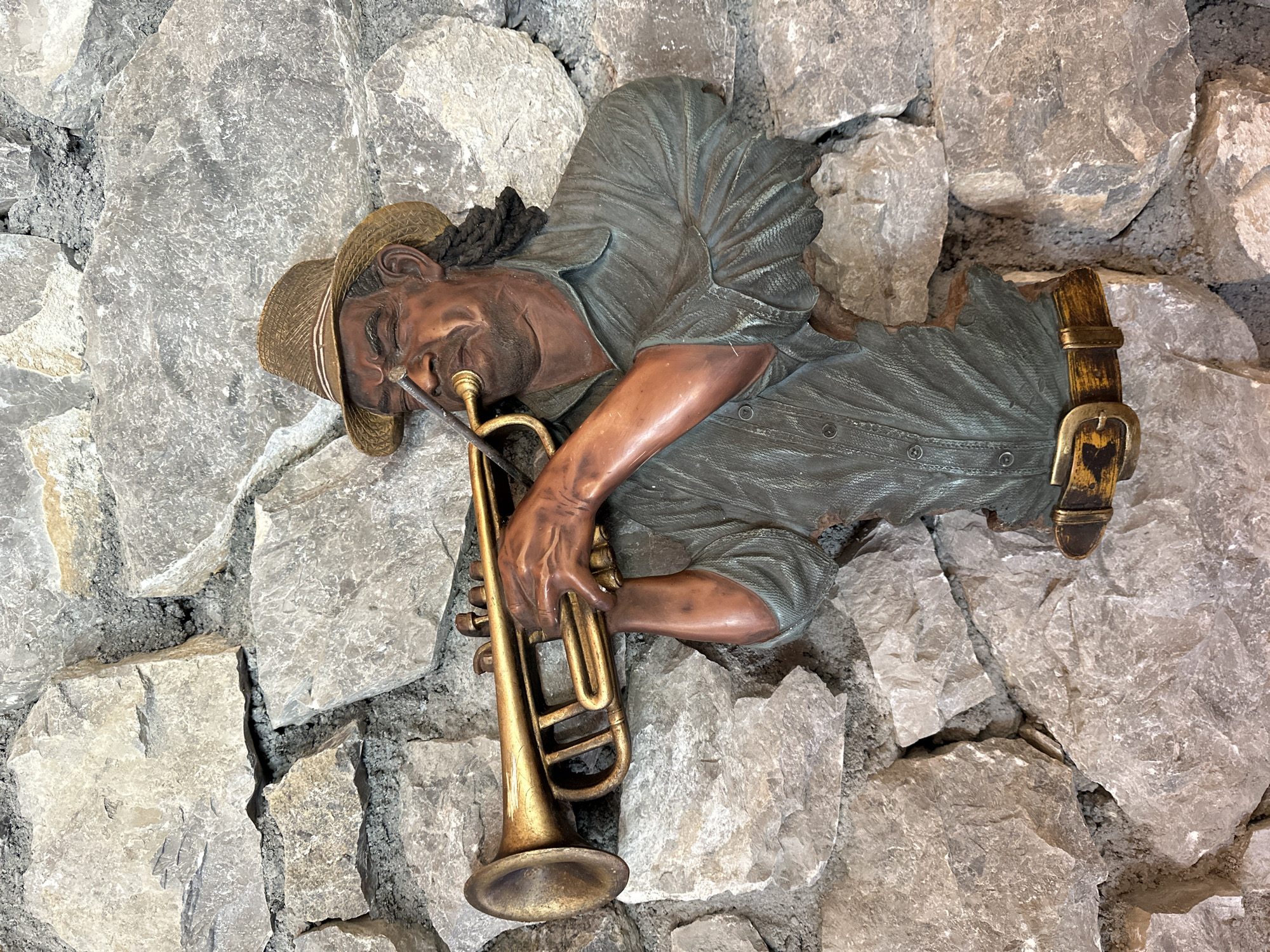 This is a medium shot photograph of a wall sculpture depicting a Black man playing a trumpet. The sculpture is integrated into a rough stone wall, with large, irregular grey rocks surrounding it. The man is wearing a straw fedora hat and a green, button-down shirt. His eyes are closed and his face is contorted as if he is deeply engrossed in playing. He is holding a golden trumpet to his lips with his right hand, while his left hand is positioned on the valves. A brown belt with a brass buckle is visible around his waist. The sculpture appears to be a relief, as only the front of the figure is visible, with the rest of his body seemingly emerging from the stone wall. The lighting is natural, casting shadows that highlight the textures of the stone and the sculpture. The location is specified as Gusinje, Montenegro.