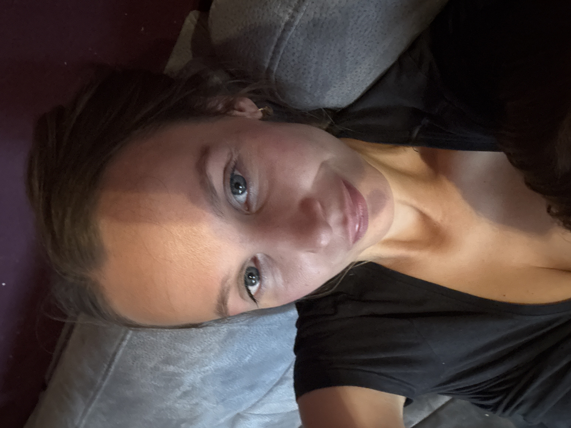 A high-angle, medium shot captures a young woman lying on her back, looking up at the camera. She has light brown hair pulled back from her face, and her blue eyes are wide open. A streak of light falls across her forehead and down the left side of her face. She is wearing a black t-shirt that is low-cut, revealing her collarbone and the top of her chest. She is wearing a small, gold hoop earring in her right ear. The background is blurry, but appears to be a gray sofa and a dark purple wall. The scene is indoors, likely in a home in Überherrn, Germany.