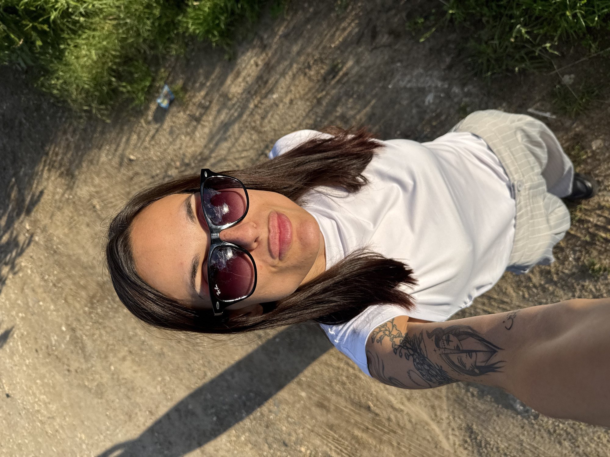 The photograph shows a woman lying on a concrete surface outdoors. She is wearing sunglasses, a white t-shirt, and light-colored trousers with a subtle stripe. A tattoo is visible on her extended arm. To the top left of the image, there is a patch of grass and some litter. The environment is bright, with shadows cast on the concrete. There is not sufficient information to place the location within the city of Pleven, Bulgaria.