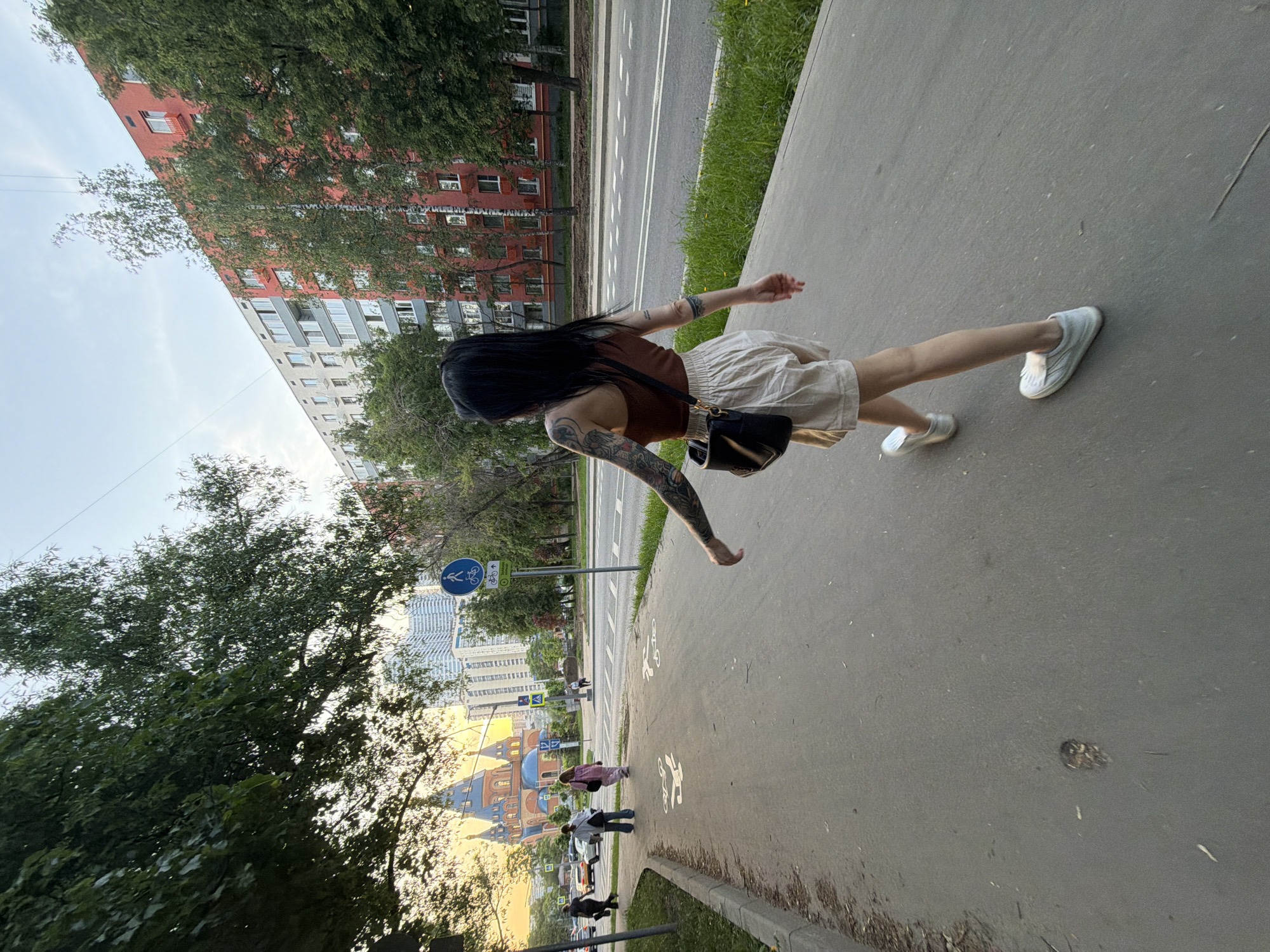 The photograph shows a woman walking on a sidewalk in Moscow, Russia. She has long black hair, a brown top, light-colored shorts, and white shoes. She carries a black purse over her shoulder. She has tattoos on her arms. The sidewalk is grey and has bicycle lane symbols painted on it. A road runs parallel to the sidewalk, with trees and buildings visible beyond. On the other side of the sidewalk, there are trees and more buildings, including one that appears to be a red and grey apartment building. A blue bicycle lane sign is visible. People are seen walking in the distance, near the brightly lit Golden Bridges.
