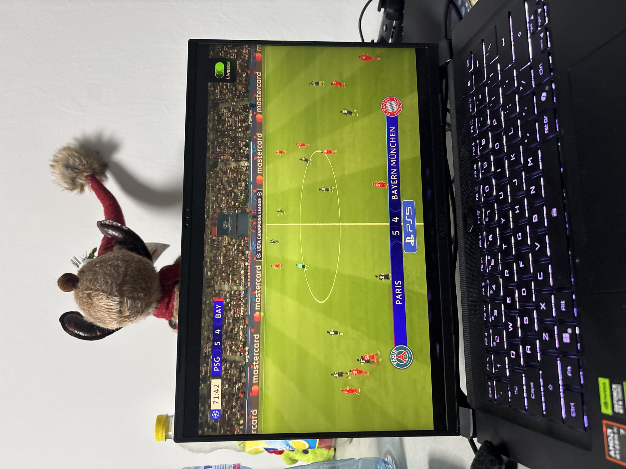 The image presents an indoor scene in Plovdiv, Bulgaria, featuring a black laptop displaying a football (soccer) game on its screen. The screen is oriented vertically, showing a match from the UEFA Champions League between PSG (Paris Saint-Germain) and BAY (Bayern München). The visible scoreboard indicates a score of PSG 5 and BAY 4, with the game time at 71:42. Both team logos (PSG and Bayern München) are clearly visible, alongside a "LiveBall" toggle and repeated "mastercard" sponsorships. A "PS5" (PlayStation) logo is also displayed on the screen. The game features players in red (Bayern) and dark uniforms (PSG) on a green pitch, with the central circle prominent.
To the left of the laptop screen, a brown, furry plush toy, resembling a reindeer or bear, with a red sweater-like garment, is positioned on a surface, casting a shadow on the white wall behind it. A plastic water bottle with a yellow cap and a colorful label is also visible beside the laptop.
The laptop's keyboard is backlit with a purple/blue glow, showing a QWERTY layout with various keys including numbers, letters, and function keys (F1-F12). Stickers on the laptop indicate "AMD Ryzen" and "GEFORCE RTX," suggesting it is a gaming laptop. Other visible text includes "AND" (partially) and various keyboard labels like "Esc," "Ctrl," "Alt," and "Fn." The overall setting appears to be a casual personal space, possibly a desk or table.