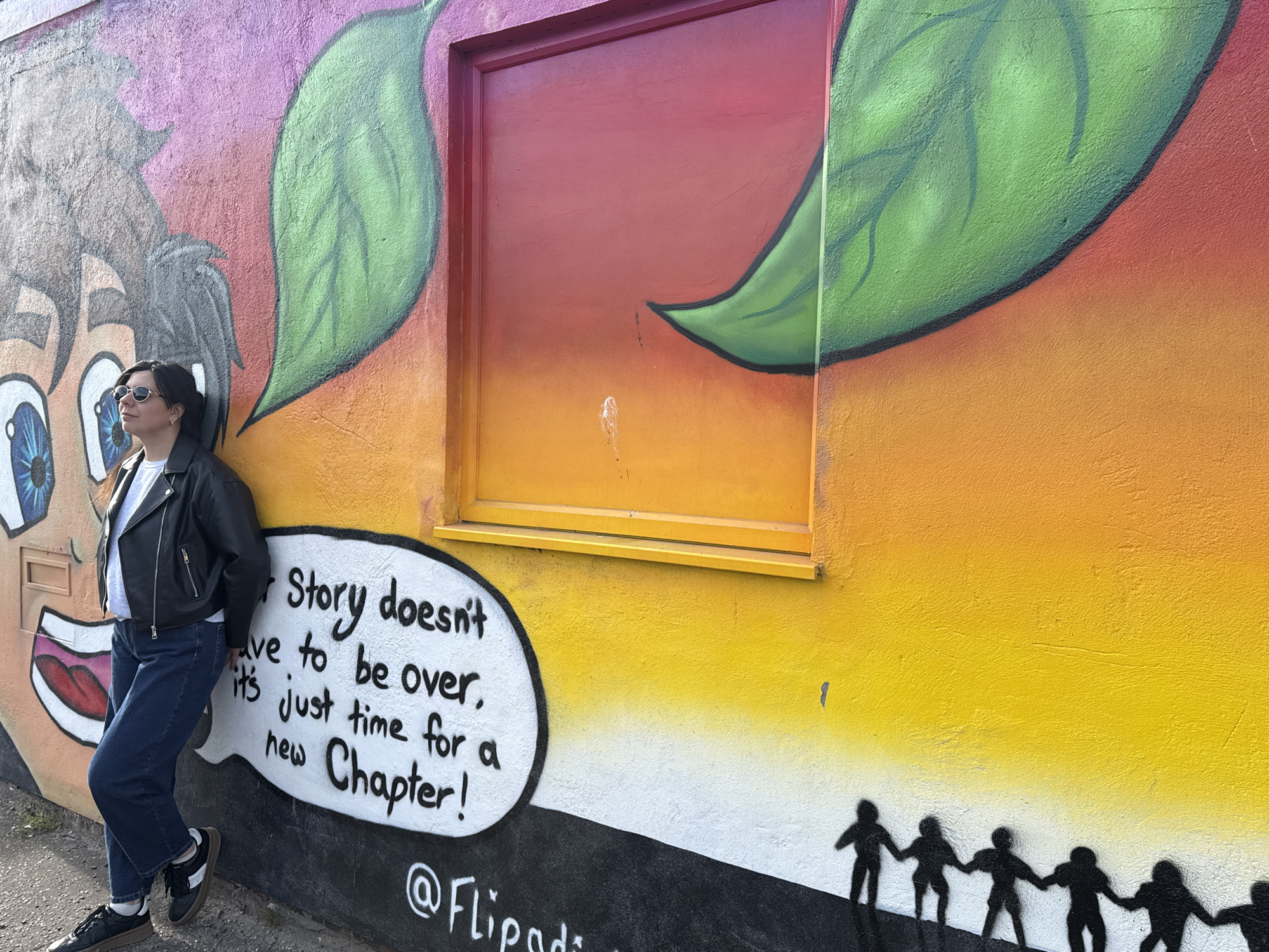A woman is standing in front of a colorful mural located in Gorleston-on-Sea, United Kingdom. The mural features a cartoon face on the left, two large green leaves, a window painted with red and orange hues, a speech bubble that reads "Story doesn't have to be over, it's just time for a new Chapter!" and at the bottom are silhouettes of people holding hands. The mural's background consists of gradient colors ranging from pink and orange at the top to yellow and white at the bottom. The woman is wearing sunglasses, a black leather jacket, a white t-shirt, and blue jeans, and is leaning against the mural.