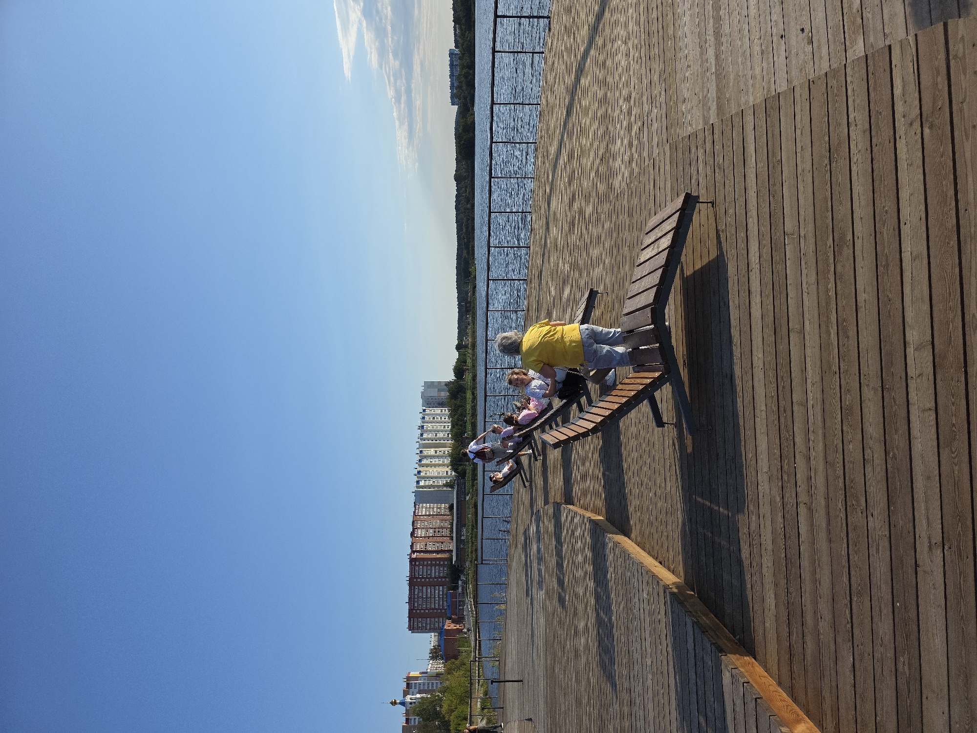 A group of people are relaxing on a wooden deck overlooking a body of water. The deck is made of weathered wooden planks and features several chaise lounges. In the background, apartment buildings line the opposite shore of the water, suggesting an urban or suburban setting. A clear blue sky with some wispy clouds suggests it is daytime. The scene appears to be located on the embankment of Lake Turgoyak in Miass, Russia, a popular recreational area.