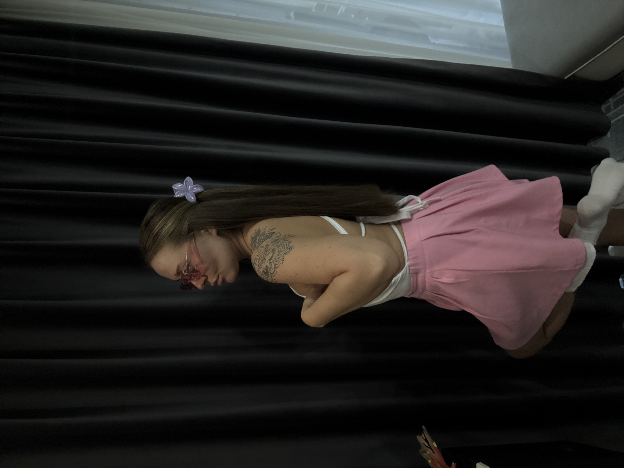 In this vertically oriented photograph, a woman is pictured from the waist up, positioned in the center of the frame. She has long, brown hair adorned with a small, purple flower accessory. She is wearing a white top and a pink skirt. A prominent tattoo is visible on her left arm. She is also wearing pink-tinted sunglasses. The background consists of dark, horizontally draped curtains, suggesting an indoor setting. To the right of the woman, her white socks are visible, and a portion of a pale-colored furniture item can be seen at the top right of the image. The lighting appears to be artificial, casting shadows that highlight the textures of the curtains. The overall mood is casual and posed. No text is visible in the image. The location is Yiwu, China.