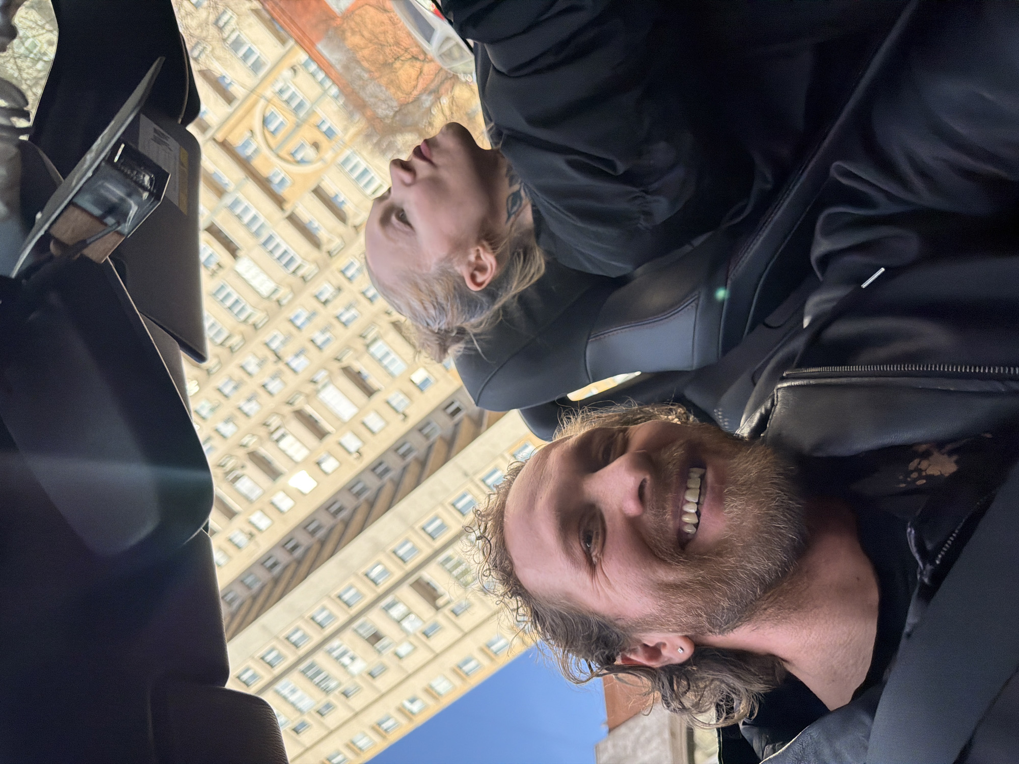 The image shows two people in a vehicle, likely a car, with a tall building visible through the windshield. The man in the foreground has curly hair, a beard, and is wearing a black jacket. He is smiling. The woman in the background has light hair and is wearing a black jacket. A tall building with numerous windows is visible outside, indicating an urban setting. Judging by the architecture of the building, it is likely located in a city, possibly Saint Petersburg, Russia. The perspective suggests the photo was taken inside the car, looking upward towards the people and the building.