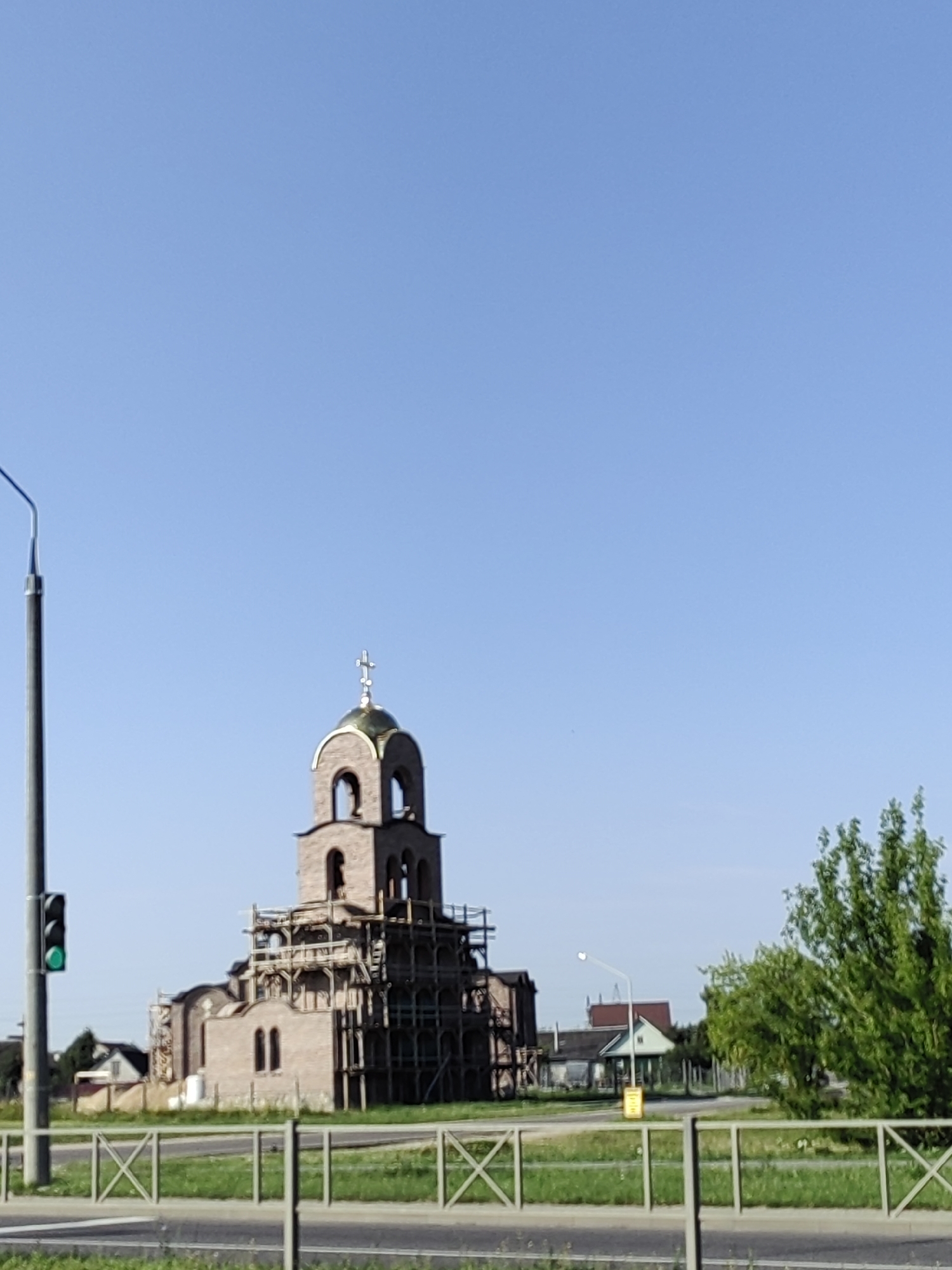 The image shows a partially constructed brick church with scaffolding surrounding it. The church has a golden dome topped with a cross. To the left of the church is a traffic light with a green signal illuminated, and a lamppost. The scene appears to be in a semi-rural or suburban area, with some houses and trees visible in the background. The sky is clear and blue. The image is taken during daylight. The location is Hrodna, Belarus.