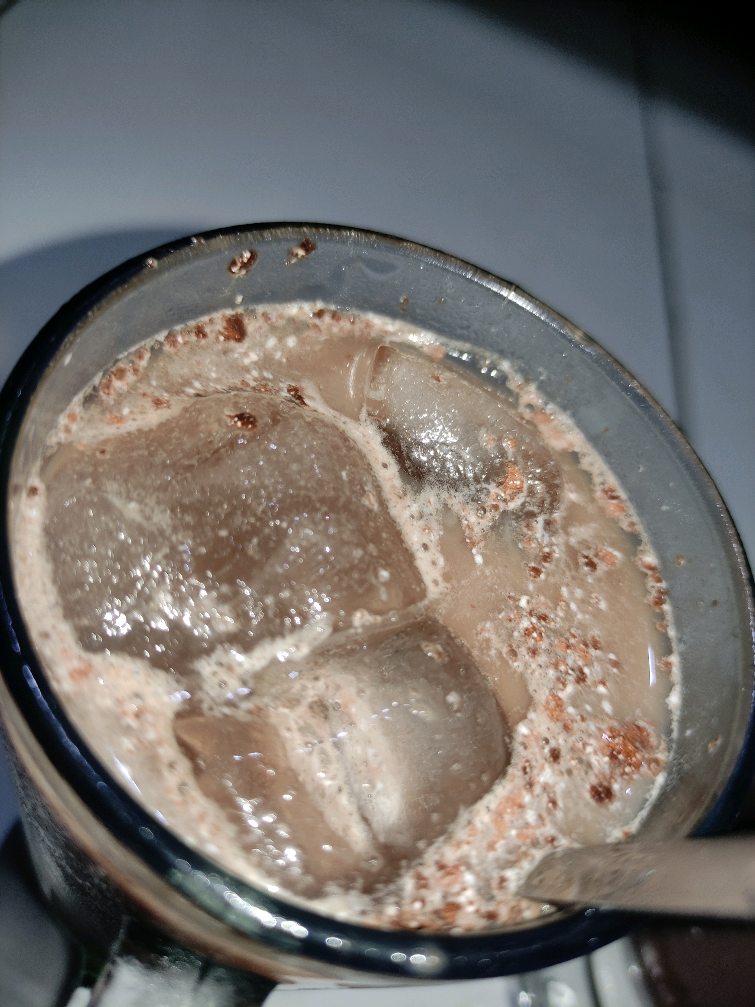 A close-up, overhead shot shows a glass of chocolate milk with ice cubes and some foam. The drink has a light brown color and appears to be slightly frothy. The ice cubes are partially submerged in the liquid, and some chocolate powder is visible on the surface and around the rim of the glass. A straw or utensil is also partially visible in the lower right portion of the frame, immersed in the drink. The background is out of focus and appears to be a neutral, light-colored surface. The image is well-lit, with reflections visible on the ice and the glass. No specific location in Ngrendeng, Indonesia can be identified from the visual information.