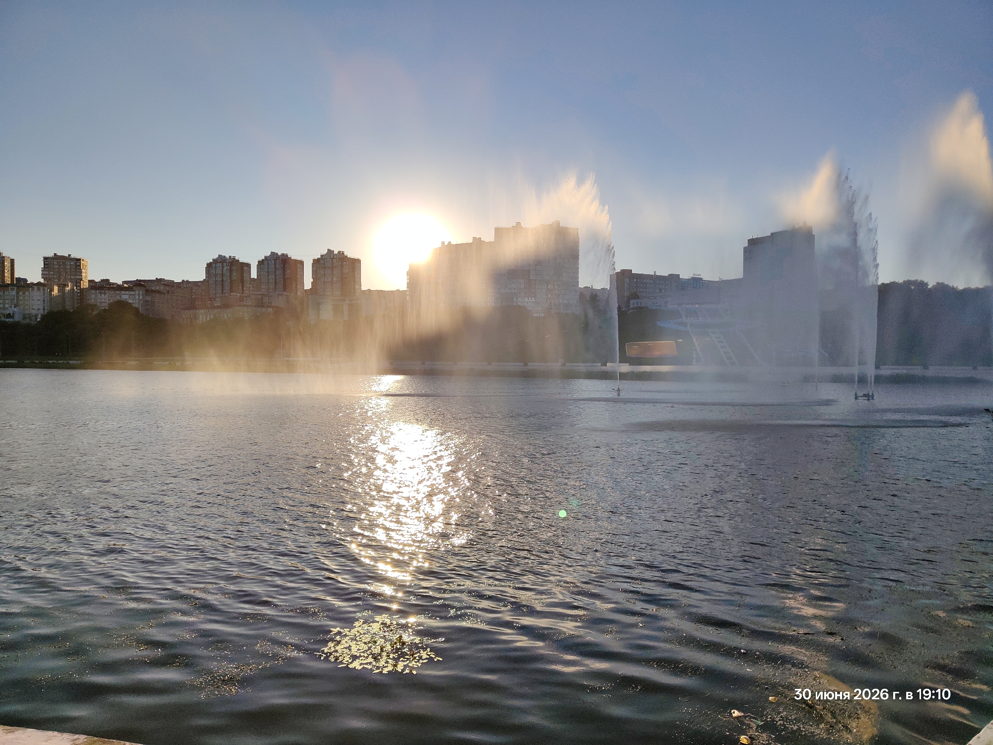 The image depicts a waterfront scene in a city with a clear sky and the sun setting or rising, casting a bright glare. A large body of water, possibly a lake or river, dominates the foreground, with gentle ripples on its surface. In the middle ground, a cityscape of apartment buildings and other structures is visible, partially obscured by mist or spray from a large fountain. The fountain itself is a prominent feature, with tall plumes of water shooting upwards, catching the sunlight. The water reflects the bright light of the sun, creating a shimmering effect. The scene conveys a peaceful and serene atmosphere, with the city's architecture juxtaposed against the natural element of the water and the man-made spectacle of the fountain. The timestamp "30 июня 2026 г. в 19:10" indicates the photo was taken on June 30, 2026, at 7:10 PM.