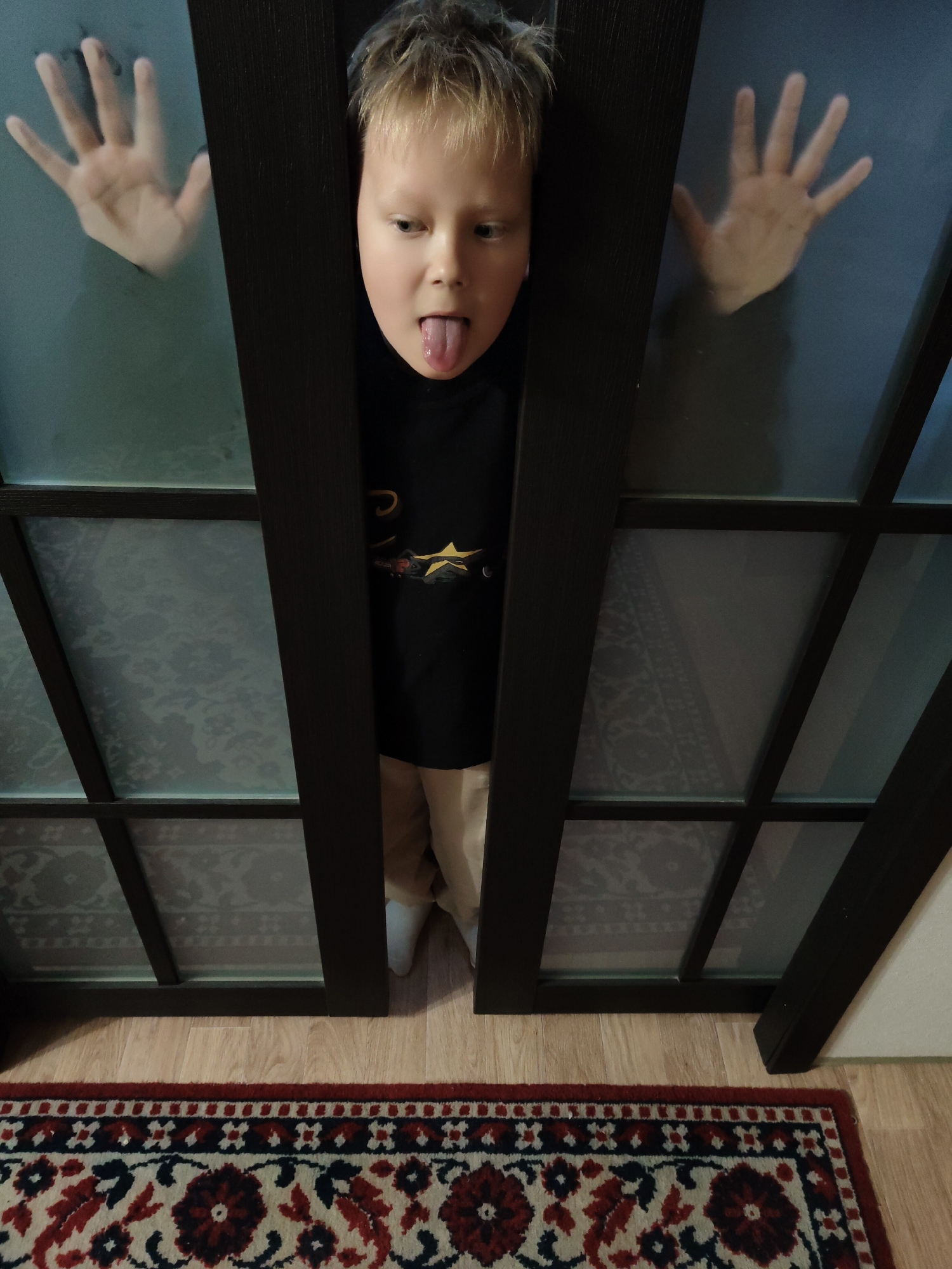 A full shot of a young boy with blond hair standing between two glass paneled doors. The boy is wearing a black shirt with a design on it, and tan pants. His tongue is sticking out. His hands are visible through the glass. Below the doors is a patterned rug on top of wooden flooring. The location appears to be indoors.