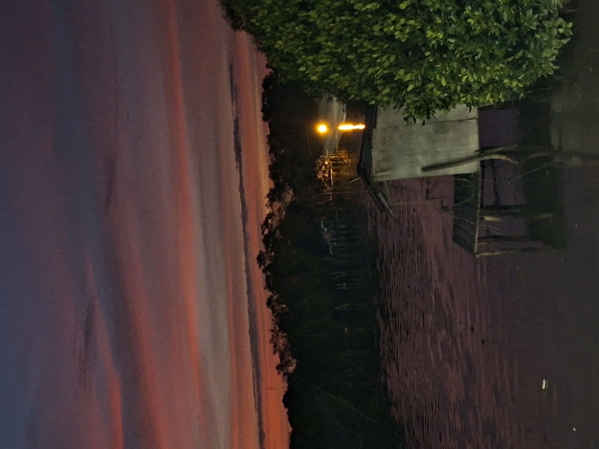 A body of water, possibly a river or bay, reflects the twilight sky which is a gradient of dark purple at the top transitioning to streaks of pink and orange closer to the horizon. A dense line of dark green trees forms a backdrop against the sky. In the foreground, a small, weathered wooden stilt house with a light-colored wall and a sloped roof sits partially submerged in the water. To the right of the house, a large, vibrant green bush with dense foliage is prominent. In the middle ground, across the water, a bright yellow light is visible, possibly from a lamp or building, casting a reflection on the water's surface. The water itself appears dark and still, with subtle ripples. The overall scene suggests a natural, possibly rural, setting at dusk in Lhokseumawe, Indonesia.