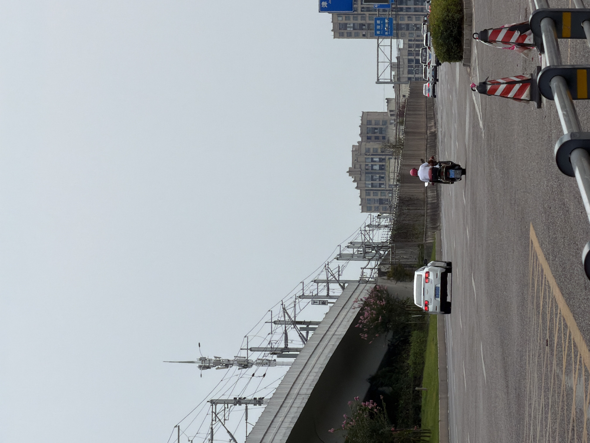 In Jiangjin, China, a two-lane road runs under an elevated overpass, likely part of a transportation network like a highway or train line, indicated by the overhead power lines and support structures. A white car with illuminated taillights is proceeding down the road. Further ahead, a person on a scooter, wearing a red helmet and white shirt, is also moving forward. Several other vehicles are visible in the distance, and traffic cones are positioned on the roadside. In the background, multi-story buildings rise against a hazy sky. The scene appears to be a typical urban street with ongoing traffic.