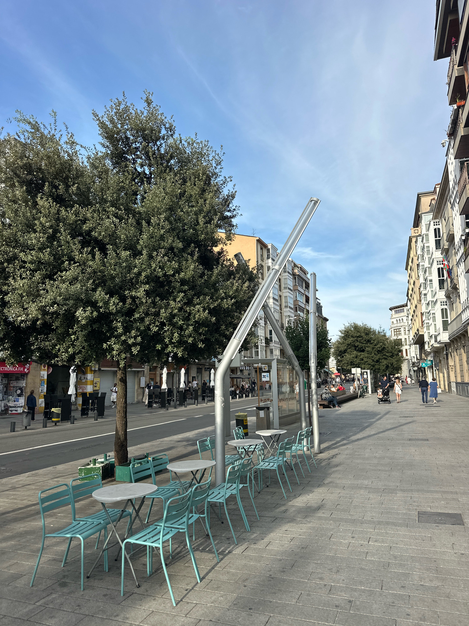 The image depicts a street scene in Vitoria-Gasteiz, Spain. In the foreground, there are several light blue metal chairs and tables set up on a paved sidewalk, suggesting an outdoor seating area for a cafe or restaurant. A large, leafy green tree stands to the left of the frame, casting a shadow over some of the seating. Further down the sidewalk, people are walking and pushing a stroller. To the right, a row of multi-story buildings with balconies lines the street, typical of urban architecture. Across the street, more buildings and some trees are visible, along with parked cars and more outdoor seating areas. The sky is blue with wispy clouds. The overall impression is a bright, sunny day in a European city.