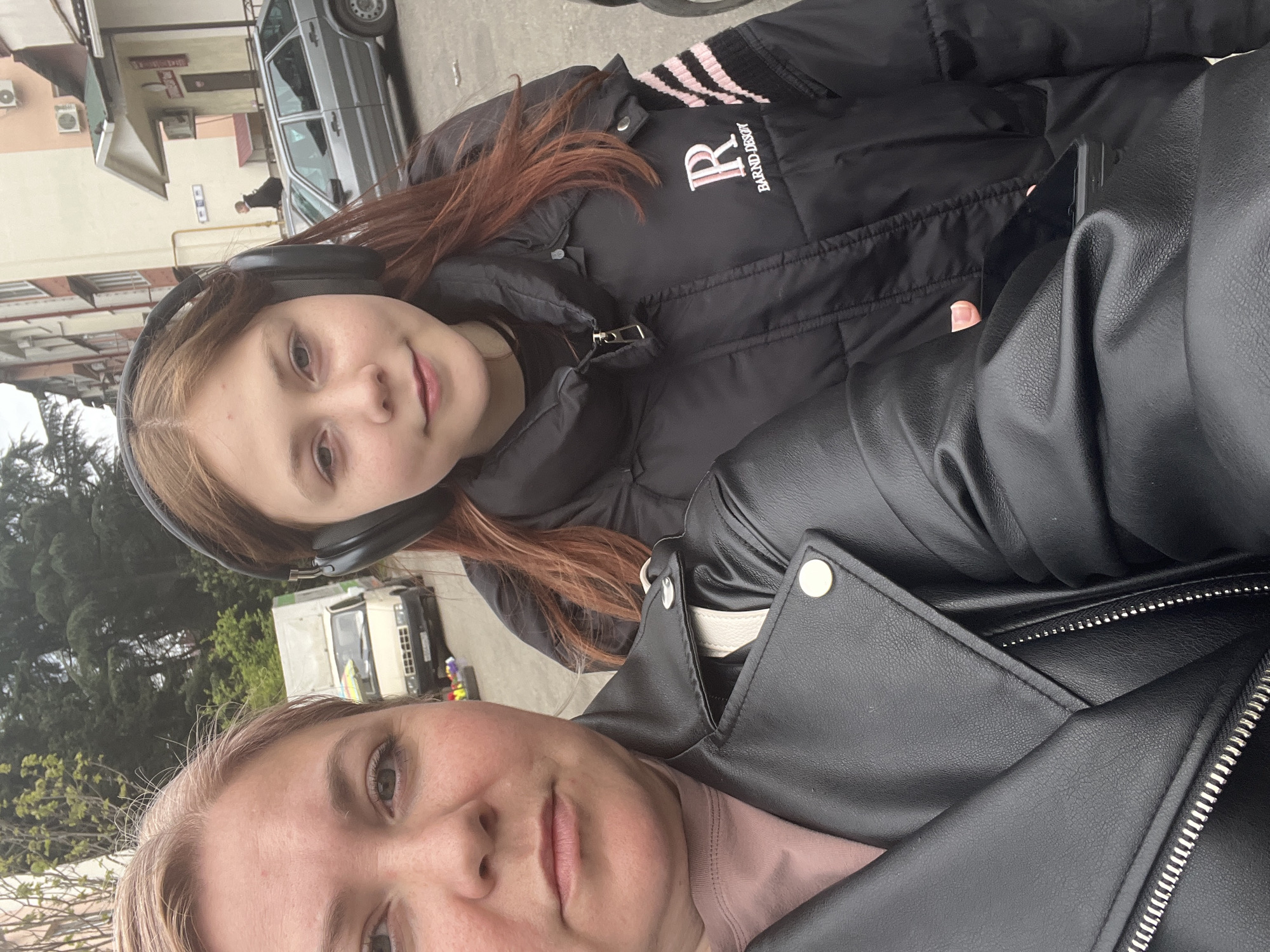 The image shows two people, possibly a mother and daughter, taking a selfie outdoors in Sochi, Russia. The daughter, positioned in the upper half of the frame, is wearing headphones and a black jacket with a logo on the chest. The mother is in the foreground, wearing a black leather jacket. Behind them, a street scene includes buildings, parked cars (including what looks like a Lada or similar Soviet-era car), and some greenery. The buildings appear to be residential or commercial structures, typical of a city environment. The lighting suggests it is daytime.