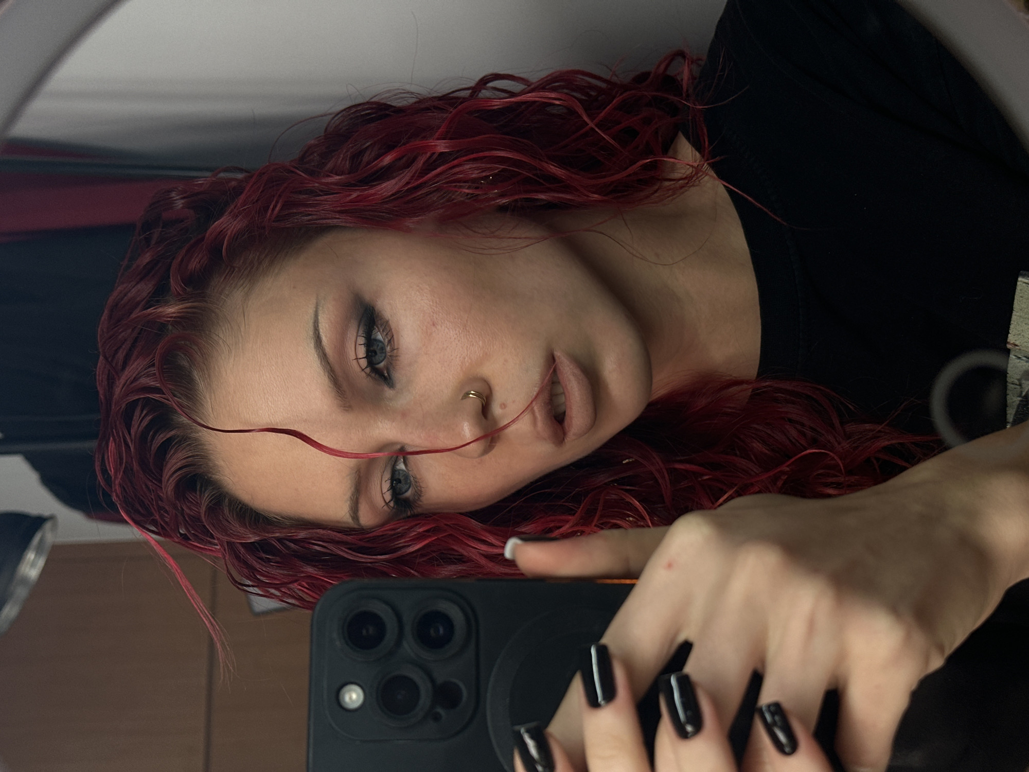 A close-up, waist-up selfie is captured in a dimly lit indoor environment. A young woman with bright red, wet, curly hair is looking at her phone, which she is holding in her left hand. She is wearing a black t-shirt. Her makeup includes dark eyeshadow and nude lipstick. Her fingernails are painted black. A gold hoop is in her nose. The background appears to be a bathroom or dressing area with a mirror reflecting the scene. No specific location in North Kuta, Indonesia is discernible from the image.