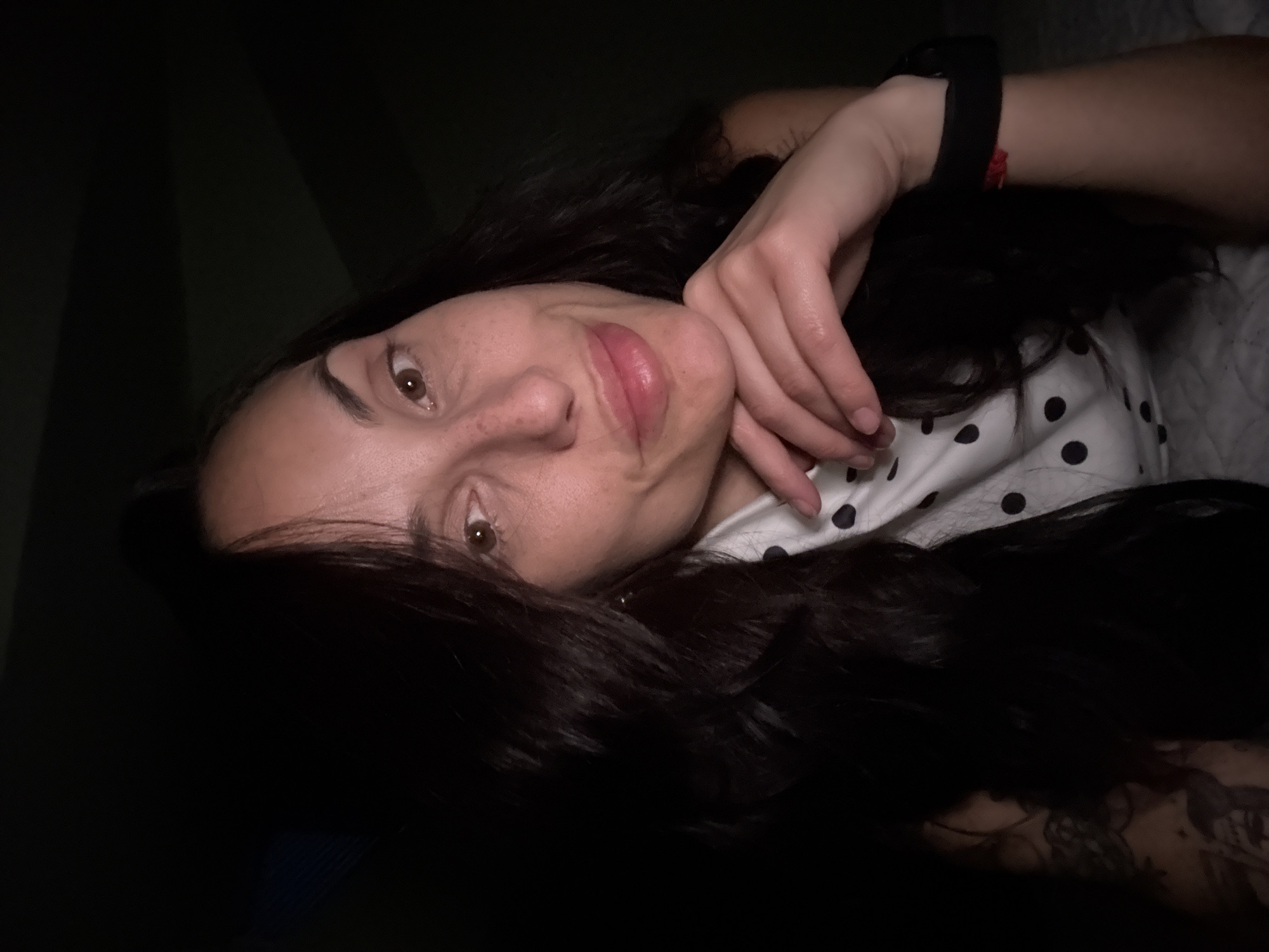 This media file presents a close-up, dimly lit portrait of a woman, possibly captured indoors in Pleven, Bulgaria. The woman is positioned with her head tilted, looking directly towards the viewer with her brown eyes. Her long, dark hair cascades around her face and shoulders. She has a fair complexion and full, pink-tinted lips, suggesting a subtle application of lip color.
She appears to be lying down or reclining, with her right hand gently resting against her cheek or chin, supporting her head. On her left wrist, a dark wristband or watch is visible. She is wearing a light-colored top, likely white, adorned with black polka dots. Portions of what appear to be intricate tattoos are visible on her left arm or shoulder, peeking out from beneath her hair and clothing.
The lighting is low, creating a soft, intimate ambiance and suggesting the photograph was taken at night or in a dimly lit room. The background is mostly dark and indistinct, with faint shadow patterns. Her expression is subtle, featuring a slight smile or smirk, conveying a relaxed and perhaps contemplative mood. There is no visible text within the image.