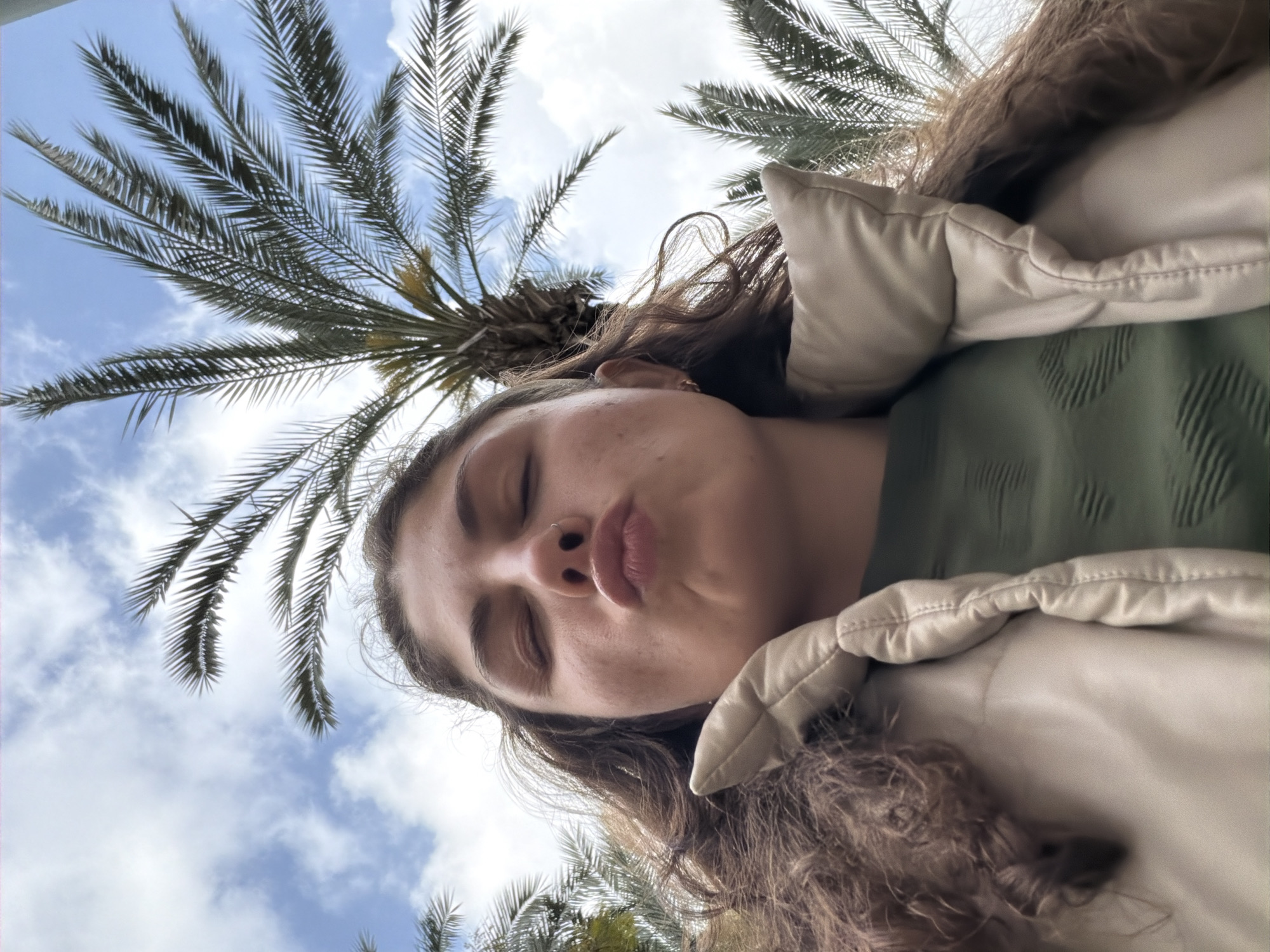 A low-angle selfie taken outdoors on a sunny day, likely in Kiryat Bialik, Israel, features a woman posing with pursed lips. Two palm trees frame the top of the shot, with a blue sky and white clouds visible between the fronds. The woman is wearing a tan jacket and an olive green shirt. Her brown hair is curly and partly obscures her face. Her eyes are closed. She is facing slightly downwards and to the side. The perspective suggests she is holding the camera above her head.