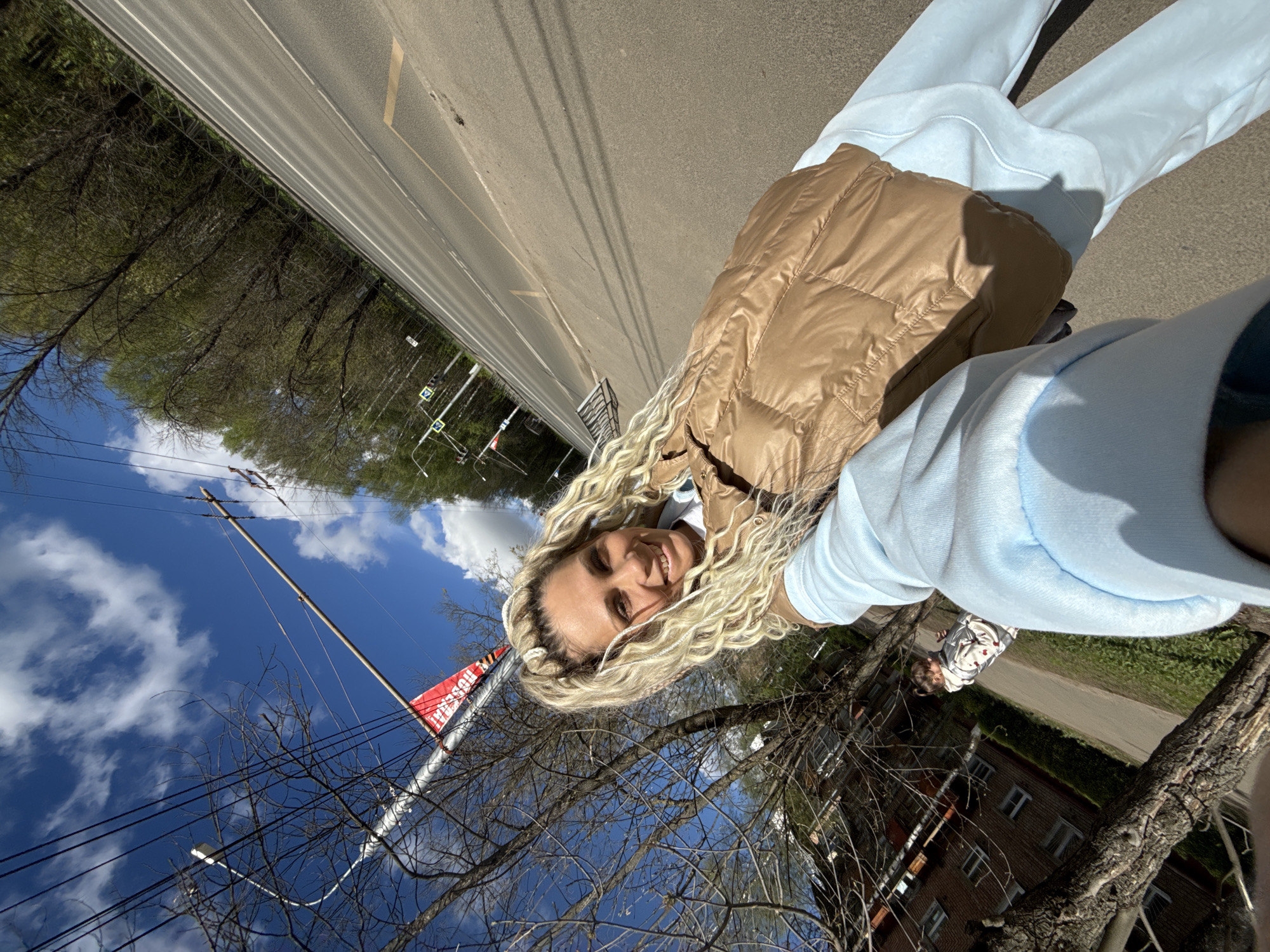 A selfie image shows a woman with long blonde, braided hair wearing a tan vest and light blue sweatshirt and sweatpants. She is smiling and looking directly at the camera. The background shows a street with white lane markings, trees, a cloudy blue sky, a building, and power lines. A sign with the word "HOSSAT" is visible near the power lines. The location is identified as the city of Шебунино, Russia.
