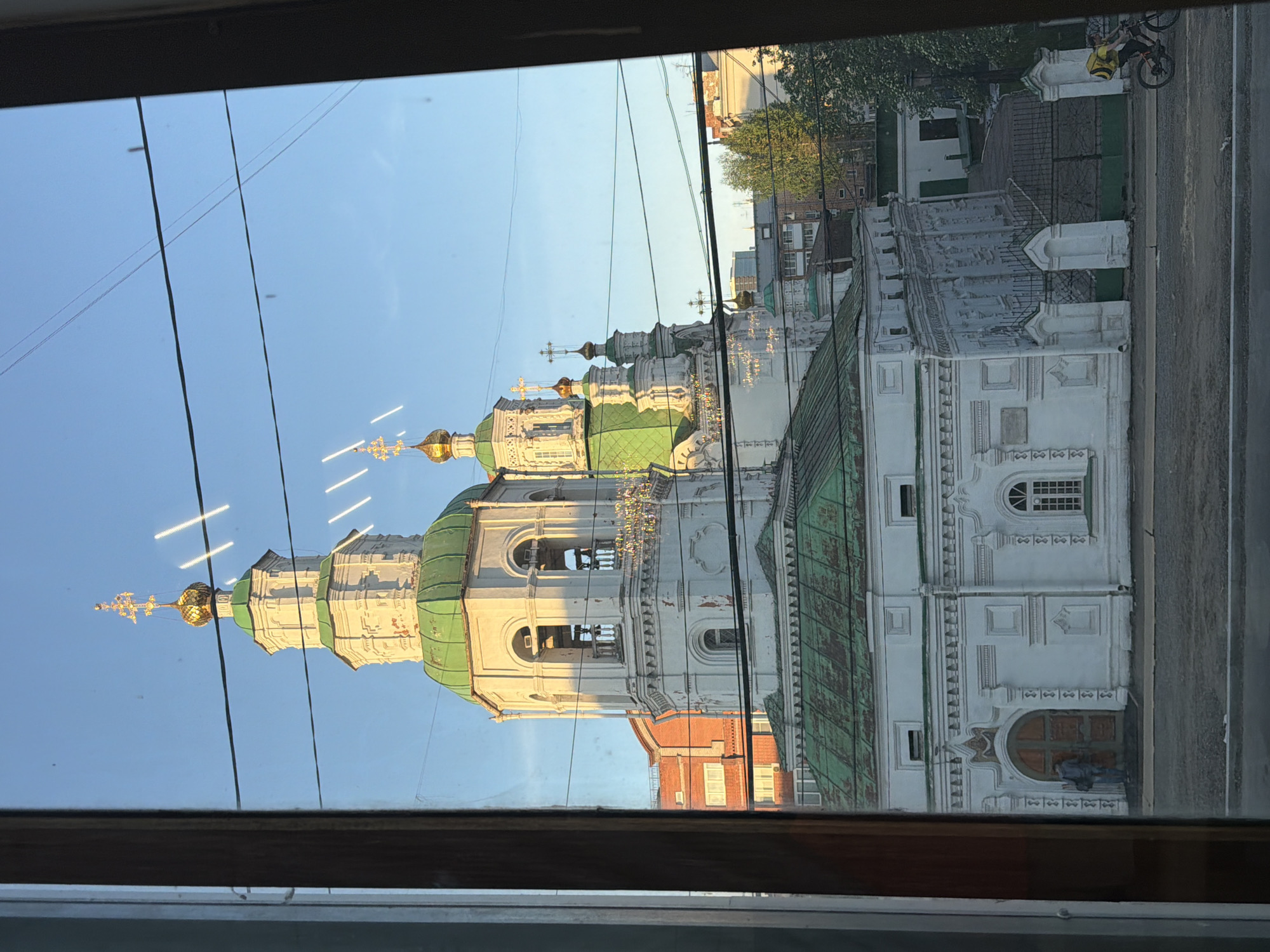 The image, viewed from what appears to be a window or opening with a dark frame, depicts an ornate Russian Orthodox church against a clear blue sky. The church is predominantly white with multiple tiers, green roofs, and several golden onion domes topped with decorative crosses. Sunlight illuminates the golden domes, suggesting a clear morning or late afternoon.
Power lines or wires are visible stretching across the blue sky in the upper part of the frame. Reflections, including bright streaks, are noticeable on the glass surface through which the photo was taken.
On the right side of the image, alongside the church, a paved street or pathway is visible. A person on a bicycle, wearing a yellow bag (possibly for delivery), is seen on this path. Another individual stands near a large, intricately carved wooden entrance door at the base of the church. In the background, behind the church, other buildings, including a red brick structure, and some green trees are visible. The scene captures an urban setting in Krasnoyarsk, Russia, under sunny weather. No visible text is present.