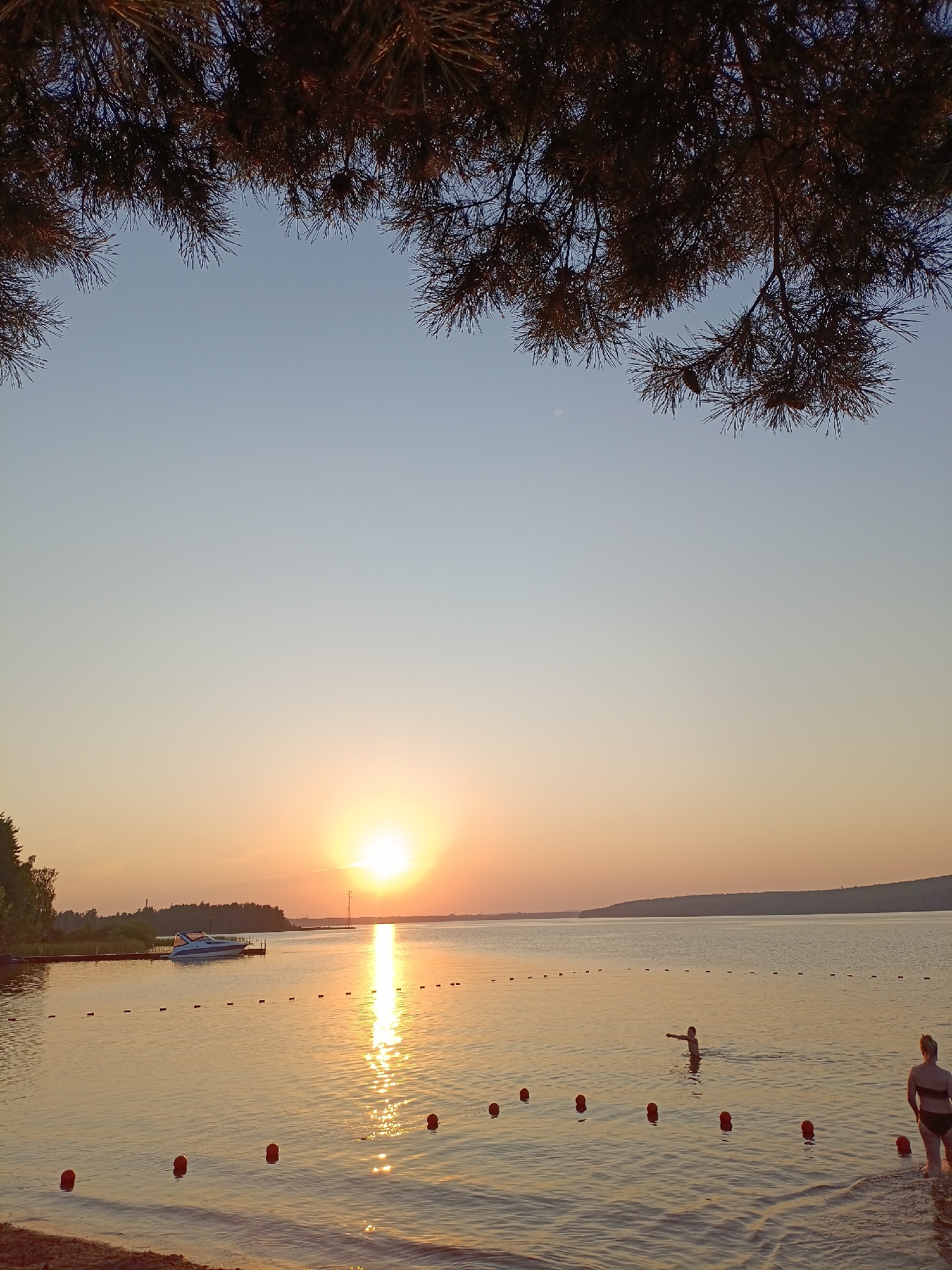The image depicts a serene sunset over the Kama River reservoir in Chaykovsky, Russia. The sun, a bright orb, is low on the horizon, casting a warm, golden glow across the water. The sky transitions from a pale blue at the top to hues of orange and yellow near the horizon. Pine branches frame the top of the image, their dark silhouettes contrasting with the bright sky.
On the water, a white motorboat is docked near a wooden pier on the left side of the frame. A string of orange buoys marks a swimming area in the foreground. Two individuals are visible in the water. One, a child, stands in the water with their arms outstretched, facing the setting sun. Another person, an adult female wearing a dark bikini, is wading out of the water on the right side of the frame, her back to the camera. The shoreline, sandy in places, is visible at the bottom left. The overall scene evokes a peaceful, end-of-day atmosphere at a lakeside or riverside recreational area.