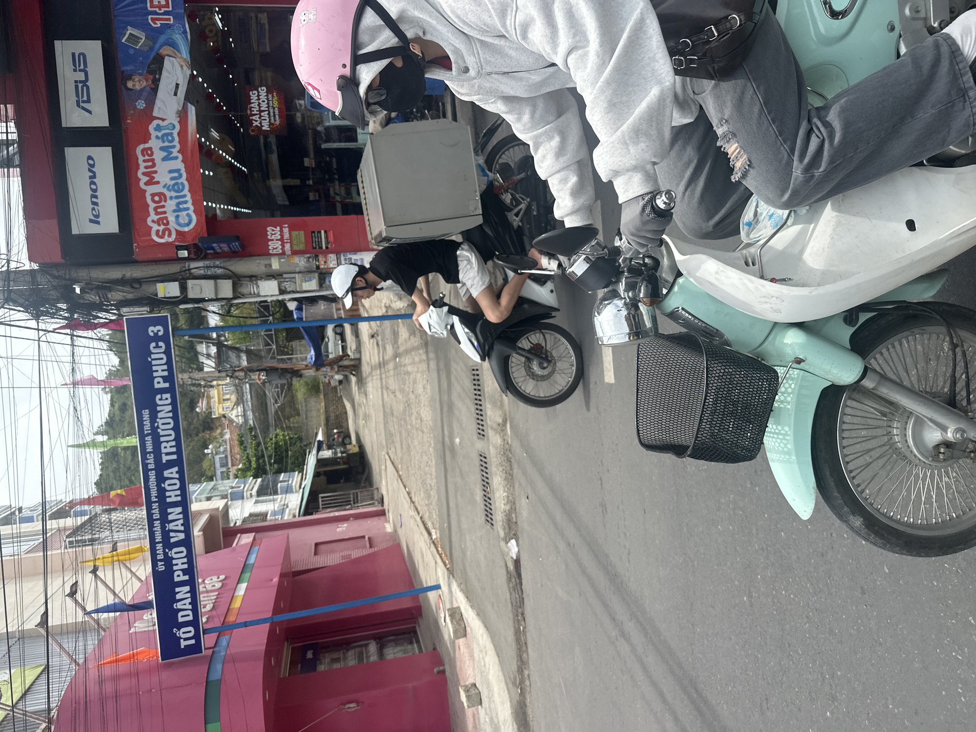 The image shows two people on motorbikes on a street in Bac Nha Trang Ward, Vietnam. The first person is wearing a pink helmet, a gray hoodie, ripped jeans, and gloves. The second person is wearing a white helmet, a black shirt, and white shorts. A blue sign reads "TỔ DÂN PHỐ VĂN HÓA TRƯỜNG PHÚC 3" indicating a cultural area. A Lenovo and Asus store is visible in the background.