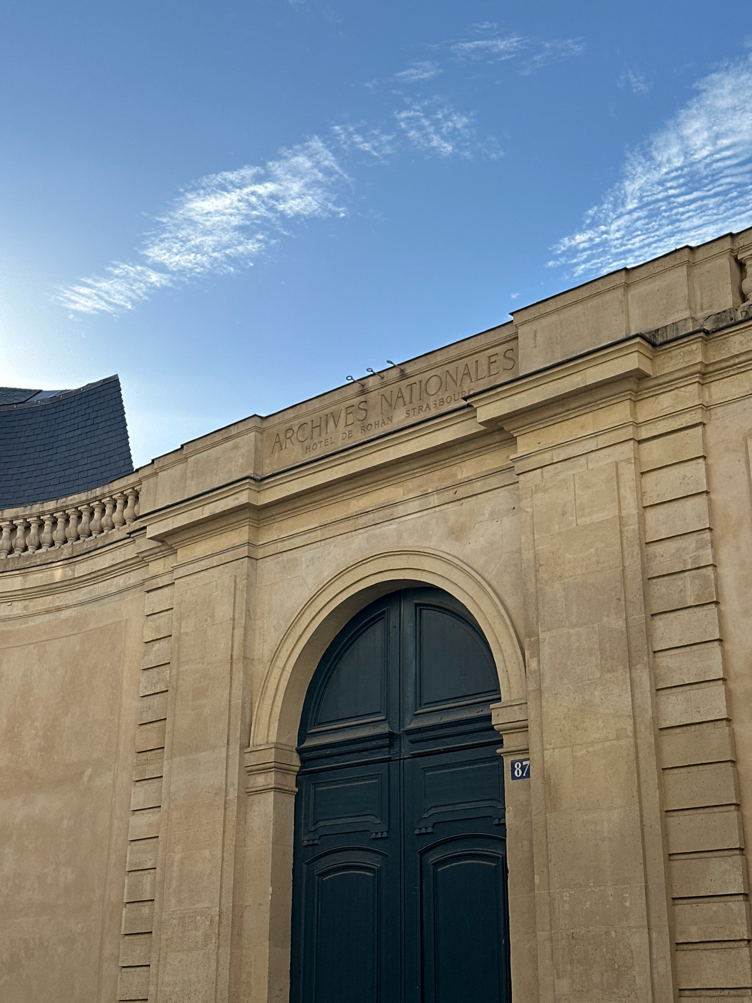 The image shows the exterior of the "Archives Nationales" at the Hotel de Rohan in Strasbourg, France. The building is made of light-colored stone, with a large arched doorway featuring dark blue double doors. Above the entrance, inscribed in the stone, are the words "ARCHIVES NATIONALES" and "HOTEL DE ROHAN STRASBOURG". A blue sign with the number "87" is visible to the right of the doorway. The sky above is blue with wispy white clouds. A balustrade and part of a dark, slate-tiled roof are visible on the left side of the building. The scene is lit by natural daylight, casting shadows on the facade. There are no people visible in the image.
