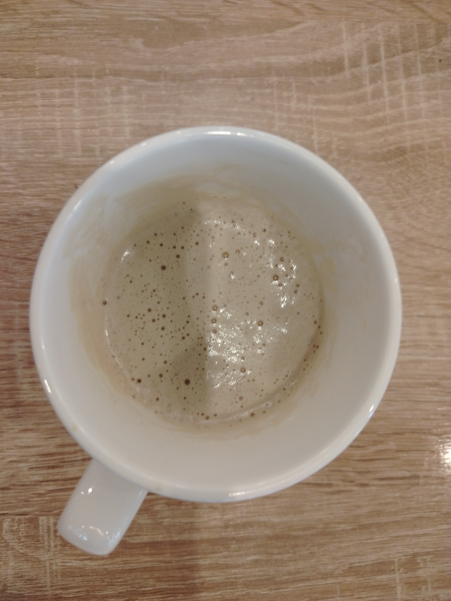 A top-down shot shows a white ceramic mug filled with frothy coffee sitting on a light brown wooden table. The coffee's surface is covered in small bubbles, and a slight residue is visible on the inside walls of the mug. The mug has a single white handle extending to the lower left of the frame. The wooden table exhibits a distinct grain pattern with visible lines and some lighter flecks. The lighting in the scene appears to be overhead, casting a subtle shadow.