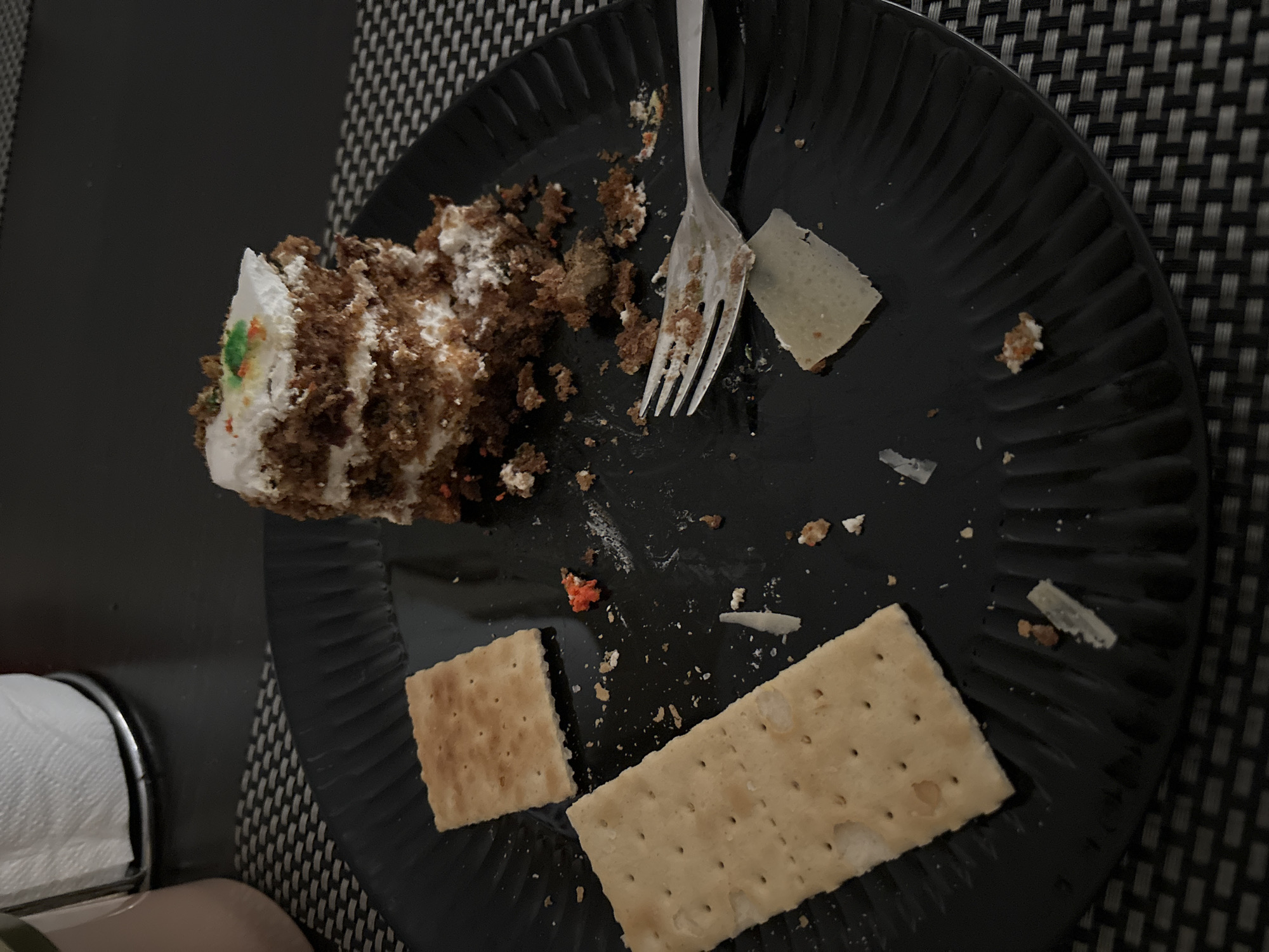 A partially eaten slice of cake sits on a black plate, next to two crackers and a fork. The cake appears to be carrot cake, with layers of cake and frosting. There are crumbs scattered around the plate, as well as some small pieces of orange and green sprinkles on the cake. The crackers are square and light brown. The fork is silver and has a few crumbs on it. The plate is round and ribbed. The background is a dark surface with a woven texture. There are no people or animals visible in the image, and there is no indication of the city of Budva, Montenegro.