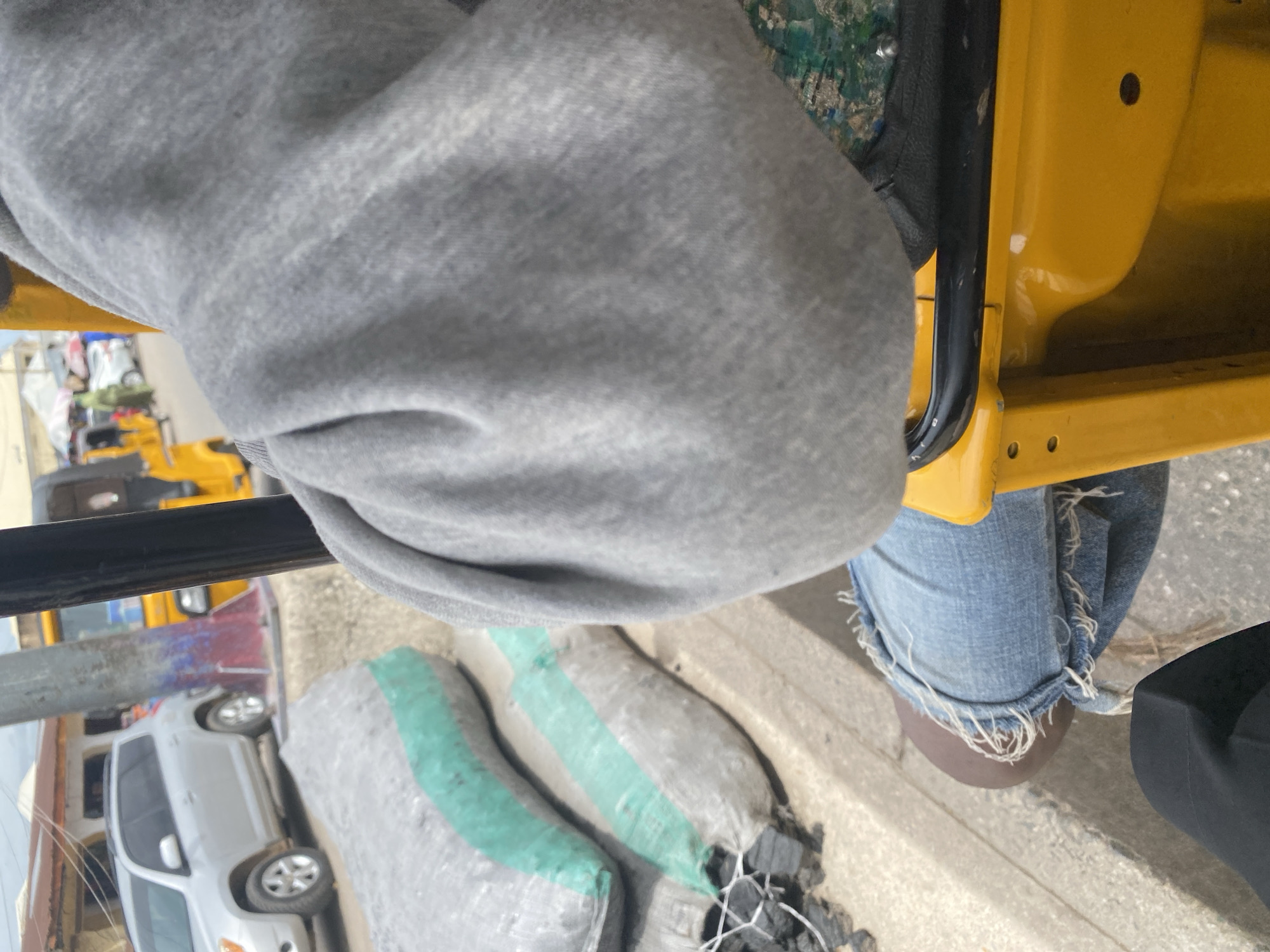 The image shows a close-up view of a person's legs and torso, wearing a gray hoodie and distressed denim jeans. The person is seated in what appears to be a yellow vehicle, possibly a tuk-tuk or a small utility vehicle, with a black handlebar visible in the foreground. The vehicle's interior is partially visible and is predominantly yellow.
Outside the vehicle, several large sacks are visible on the ground. These sacks appear to contain charcoal, with some green netting visible on two of them. The sacks are dark gray and bulging. In the background, a light-colored SUV is parked, suggesting an urban or semi-urban environment. Other vehicles and structures are also faintly visible, hinting at a bustling street scene.
The lighting suggests it is daytime, with no specific indicators of the time of day or weather conditions. The overall impression is that of someone transporting goods, possibly charcoal, in a busy area. The focus on the person's clothing and the cargo suggests a candid or street photography style. The location context provided is Jalingo, Nigeria. There is no visible text in the image.