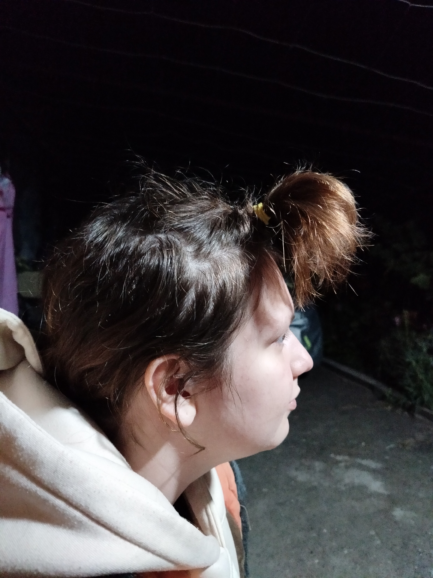 A side profile of a person's head and shoulders is captured in a close-up, outdoor shot. The person is wearing a light-colored hooded garment. Their hair is dark brown and appears somewhat disheveled, with a small portion of it pulled into a bun on top of their head, secured with a yellow elastic band. The background is dark and out of focus, suggesting it is nighttime. The environment appears to be a paved area with some foliage visible in the background. The image is taken at an unknown location in Unknown, Unknown.