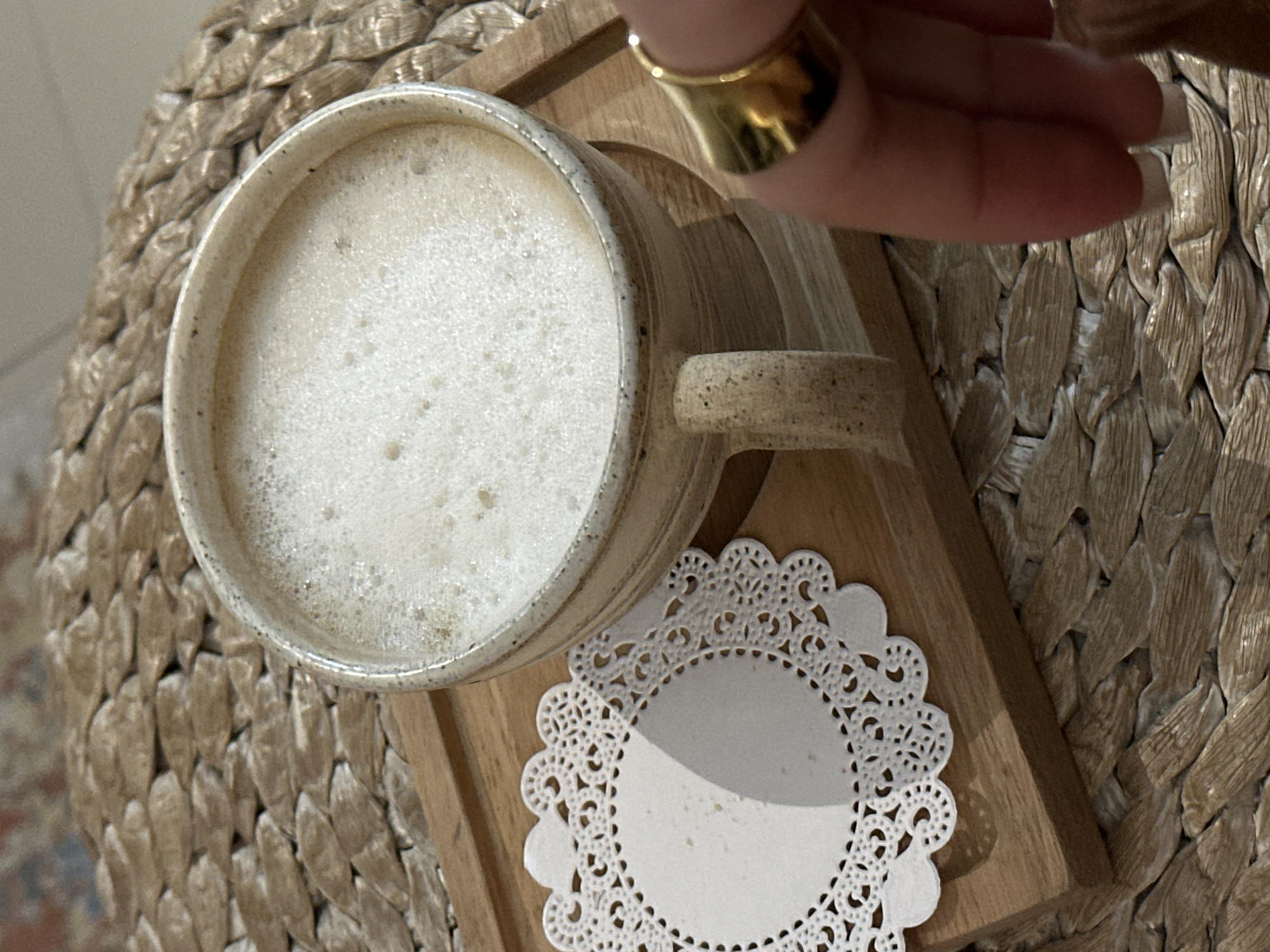 A high-angle shot shows a mug filled with a light-colored, frothy beverage, likely coffee or latte, sitting on a small wooden tray with a white doily underneath. A hand wearing a wide gold ring is partially visible in the upper right corner of the frame. The setting appears to be a cafe or home in Kerobokan Kelod, Indonesia, based on the background texture that resembles woven straw or rattan, common in the area. The objects are stationary, and there is no apparent interaction between the hand and the mug. The environment seems to be well-lit, suggesting daytime.