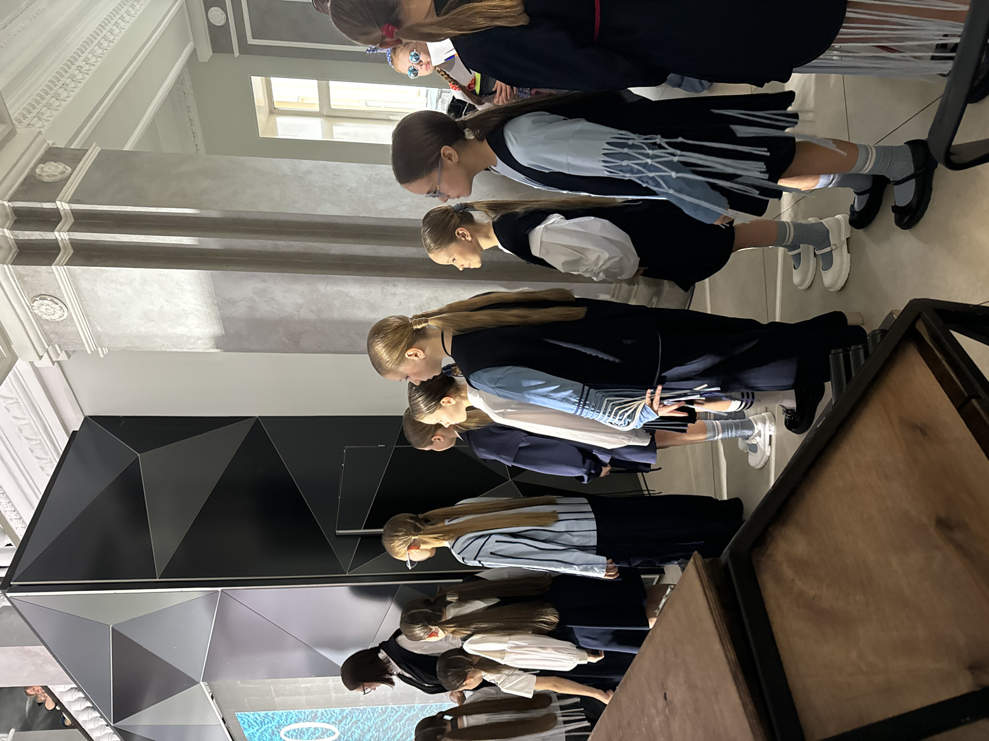 A line of girls is standing in front of a geometric wall in what appears to be an exhibition or performance space. The location is Krasnoyarsk, Russia.
The girls are wearing school uniforms with varying designs. Their hair is pulled back into ponytails or similar styles. Most of them are fair-skinned. Their heads are tilted down.
The wall has a geometric pattern made of triangular and polygonal shapes in shades of black, gray, and white. There is a monitor or display screen at the bottom of the wall.
The space has an industrial or modern architectural design with high ceilings, ornamental detailing, and a beam structure. A wooden table or display fixture is visible. Natural light comes from a window.