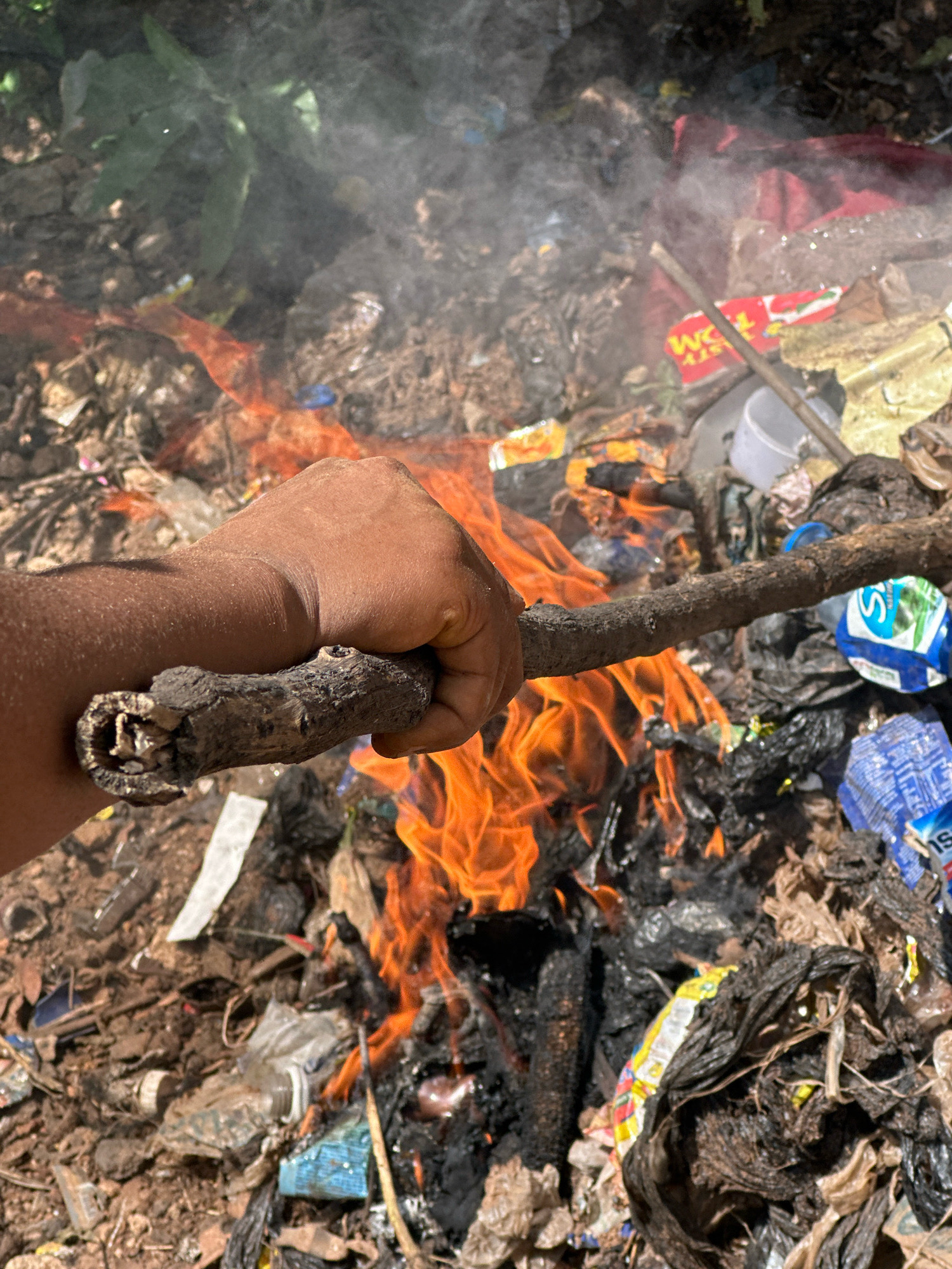 A close-up, ground-level shot shows a hand holding a thick, gnarled stick over a pile of burning garbage. The hand, exhibiting a dark skin tone, is clenched around the stick, which has a rough, bark-like texture and appears to be dry and brittle. In the background, the garbage heap is consumed by vibrant orange flames and wisps of grey smoke, creating a chaotic scene. The refuse consists of a variety of materials, including plastic bags, bottles, paper packaging, and what appears to be fabric or cloth. The overall environment is one of disarray and pollution, with the burning debris suggesting an informal waste disposal method. The context of Gura Top, Nigeria, indicates a specific urban or peri-urban setting where such practices might be common due to inadequate waste management infrastructure.