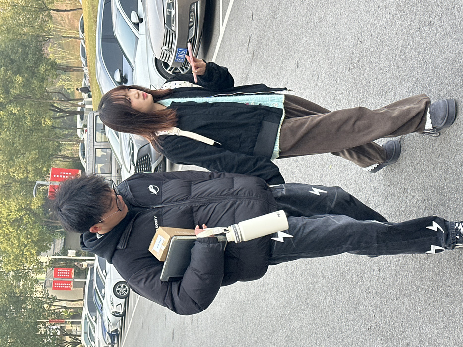 Two young people stand on a gray surface, likely pavement. The person in the foreground wears a black puffer jacket and black jeans adorned with white lightning bolts. They hold a book, a small package, and a white water bottle. The person behind them wears a black jacket over a light blue sweater, brown pants, and is holding up a pink phone, possibly taking a picture. Cars are parked in the background along with some trees.