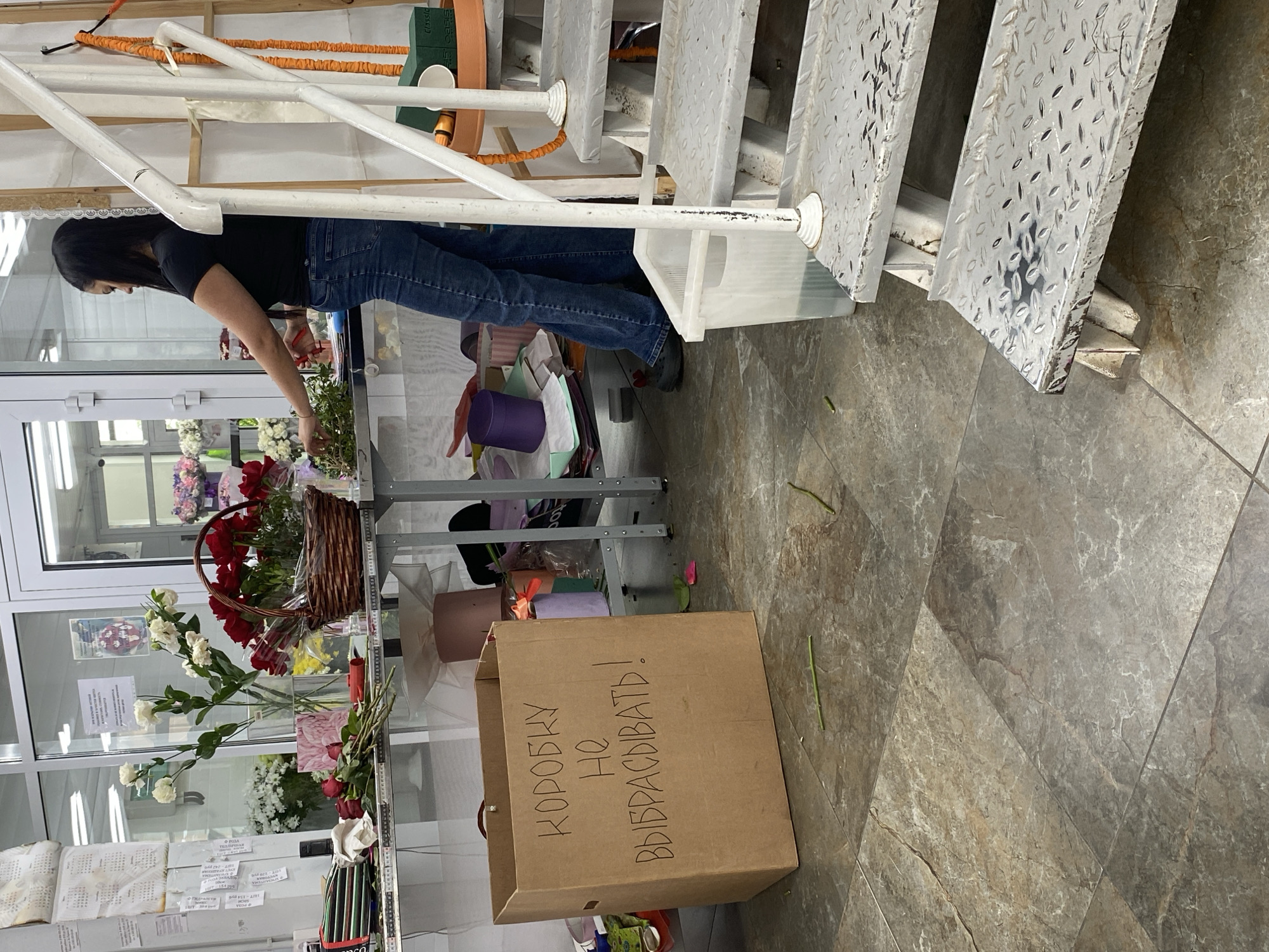 A woman with dark hair is arranging flowers in a shop. She is wearing jeans and a black top, bending over a display. The shop's interior has white shelves filled with flowers, vases, and arrangements. A cardboard box on the floor has writing in Russian. The floor is tiled. Stairs are visible in the background. The location is likely Yekaterinburg, Russia, given the writing on the box.