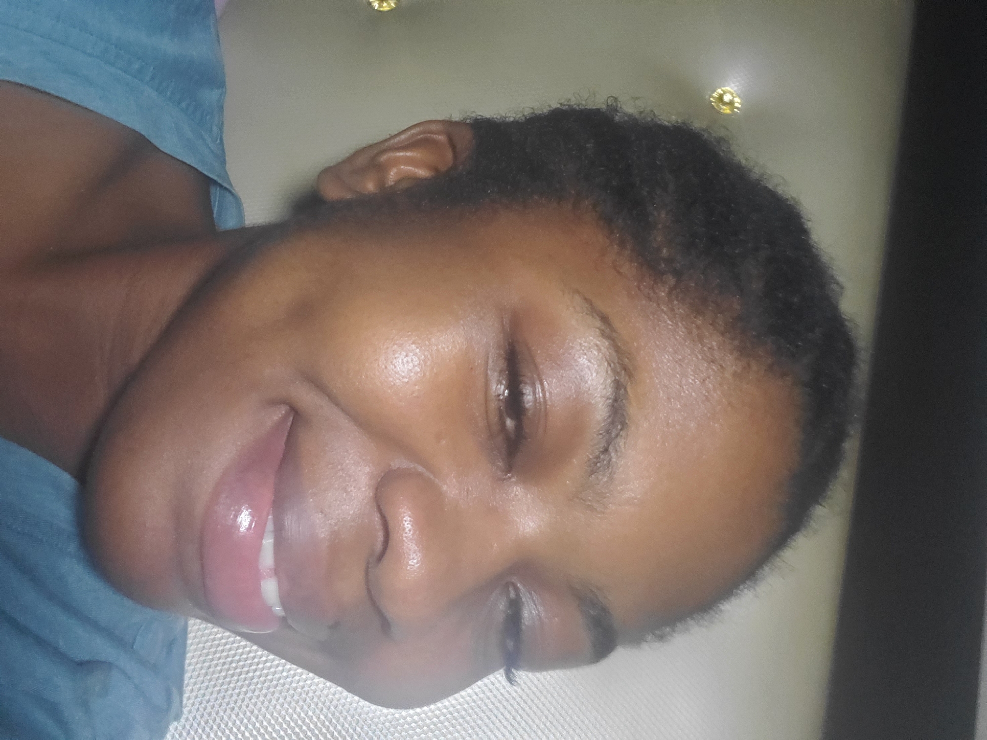 A close-up portrait features a woman with dark skin and short, dark, textured hair, centered in the frame. She is smiling broadly, with her eyes partially closed in a squint, revealing the top row of her teeth. Her lips appear full and are covered with a glossy substance, reflecting light. A small, circular, yellowish stud earring is visible in her left ear. She is wearing a blue garment, likely a shirt or top, visible around her shoulders and chest. The background is out of focus, showing light-colored, possibly textured panels. On the left side of the image, behind her head, a round, shiny, yellowish-gold decorative stud or button is visible against a muted background. No identifying features regarding the city of Kakau, Nigeria, are discernible within the image.