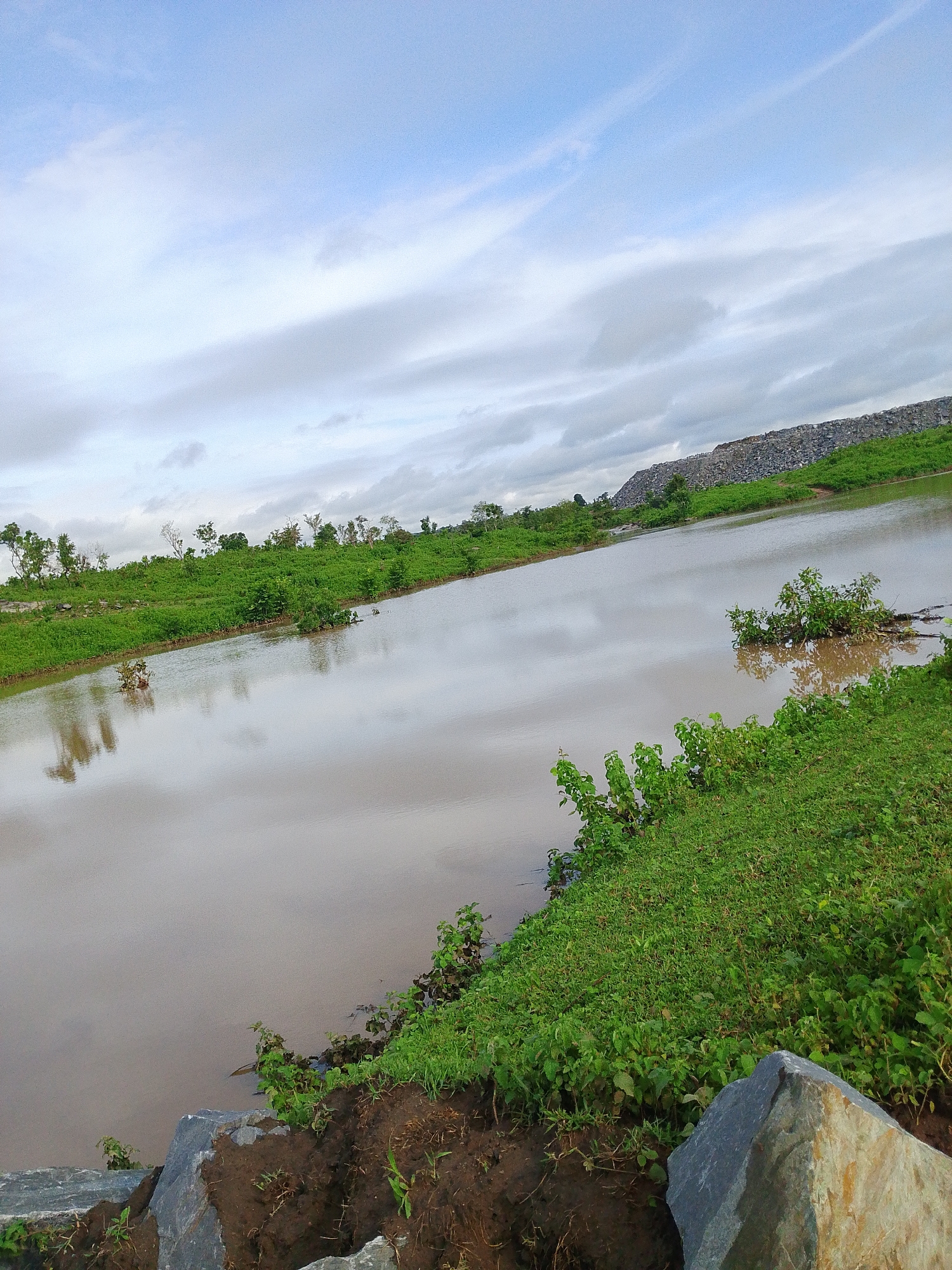 The image shows a body of muddy water with reflections of the sky and surrounding vegetation. The water is calm, with some sparse green plants emerging from it. The foreground features the muddy bank of the water with large grey rocks. Beyond the water, there is a lush green landscape with various trees and shrubs. In the background, a large pile of rubble or debris is visible, contrasting with the natural environment. The sky is partly cloudy with a mixture of blue and white. The overall scene depicts a natural setting near a body of water, with the presence of man-made debris suggesting a nearby industrial or waste disposal area. The location is stated to be Indo, Nigeria.
