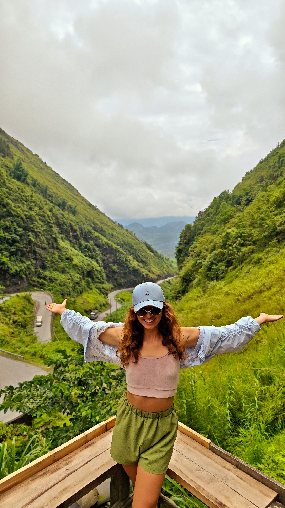 A woman stands on a wooden platform with her arms outstretched, smiling at the camera. She wears a blue baseball cap, sunglasses, a pink crop top, a light blue unbuttoned shirt, and green shorts. The background features a valley with lush green mountains and a winding road. The sky is cloudy. This is likely in the Phố Bảng Commune, Vietnam, given the landscape.