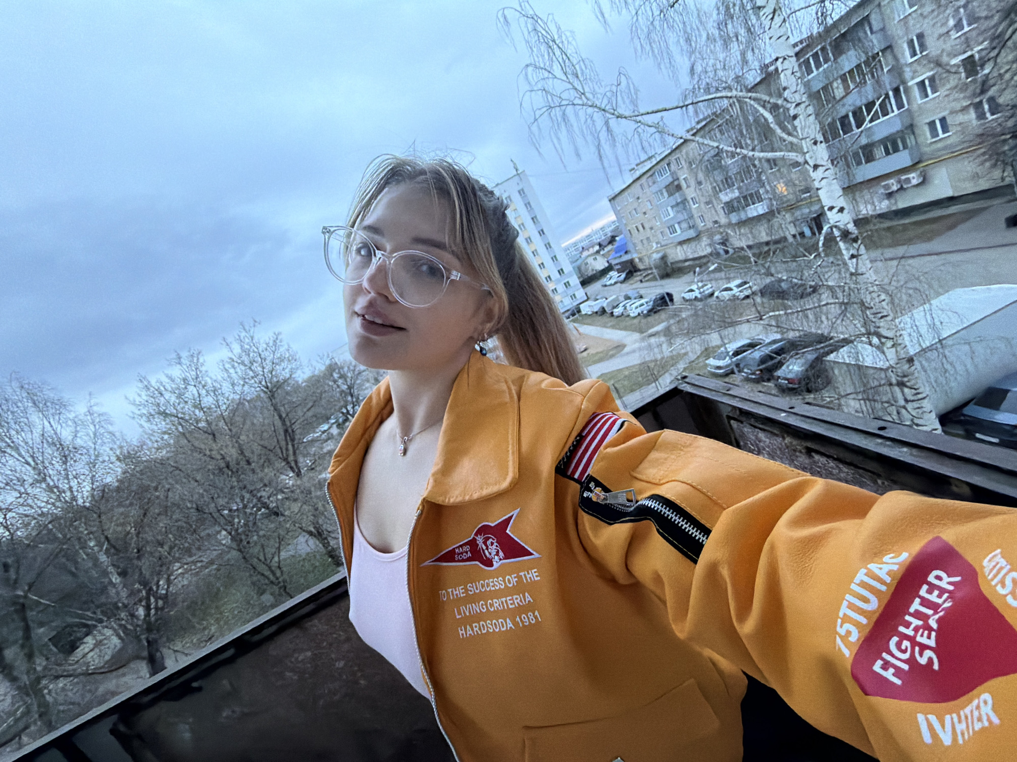 A woman in glasses and a yellow jacket is taking a selfie on what appears to be a rooftop. In the background, there are apartment buildings, trees, and parked cars, suggesting an urban environment. The jacket has some writing and symbols on it, including "HARDSODA 1981," "FIGHTER SEAS," and "15TUTAC IVHTER". The overcast sky and bare trees suggest it might be late autumn or early spring. Based on the visual elements, it's plausible this image could be taken in Naberezhnye Chelny, Russia, as the architecture and urban layout align with the city's characteristics.
