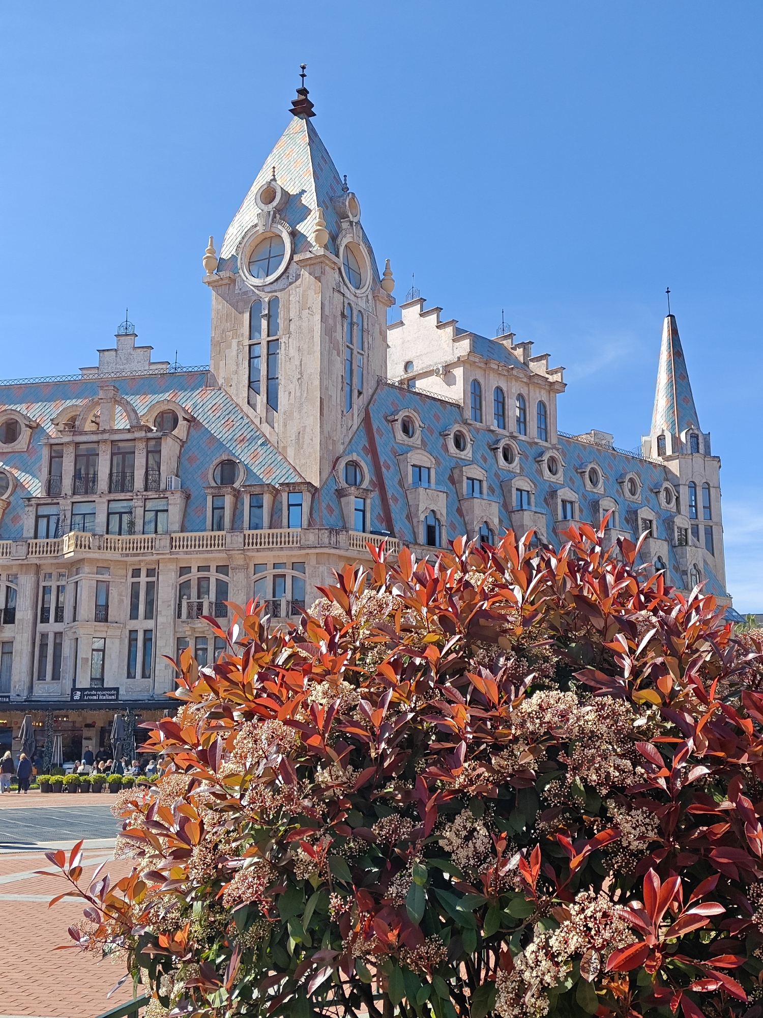 The image shows a building in Batumi, Georgia, with a distinct architectural style featuring a tower, a steeply pitched roof with decorative tiles, and numerous windows. The building is likely a hotel or a public building in a central location. In the foreground is a large bush or shrub with red and green leaves and small white flowers. The sky is clear and blue. The lower-left corner of the picture contains people walking in the background. The building is light-colored with blue and red roof decorations.