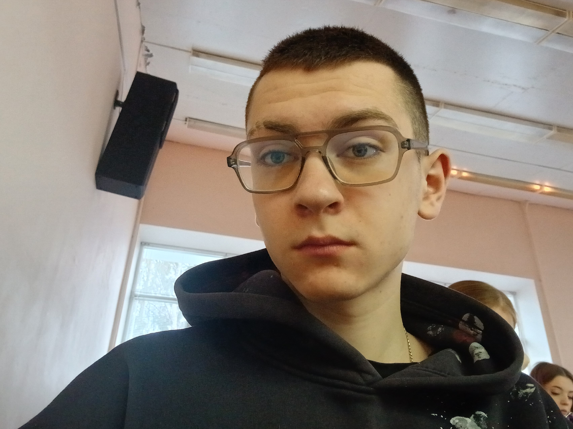 A close-up shot features a young man with short, dark hair and light eyes, wearing light brown-rimmed glasses and a black hoodie. A gold chain is visible around his neck. His expression is neutral. A black rectangular speaker is mounted on the light-colored wall in the upper left corner. A window is visible in the background. Portions of other individuals are visible in the background. The setting suggests an indoor environment, possibly a classroom or public building, based on the fluorescent lighting, and window. The location of Chaikovsky, Russia is not readily apparent.